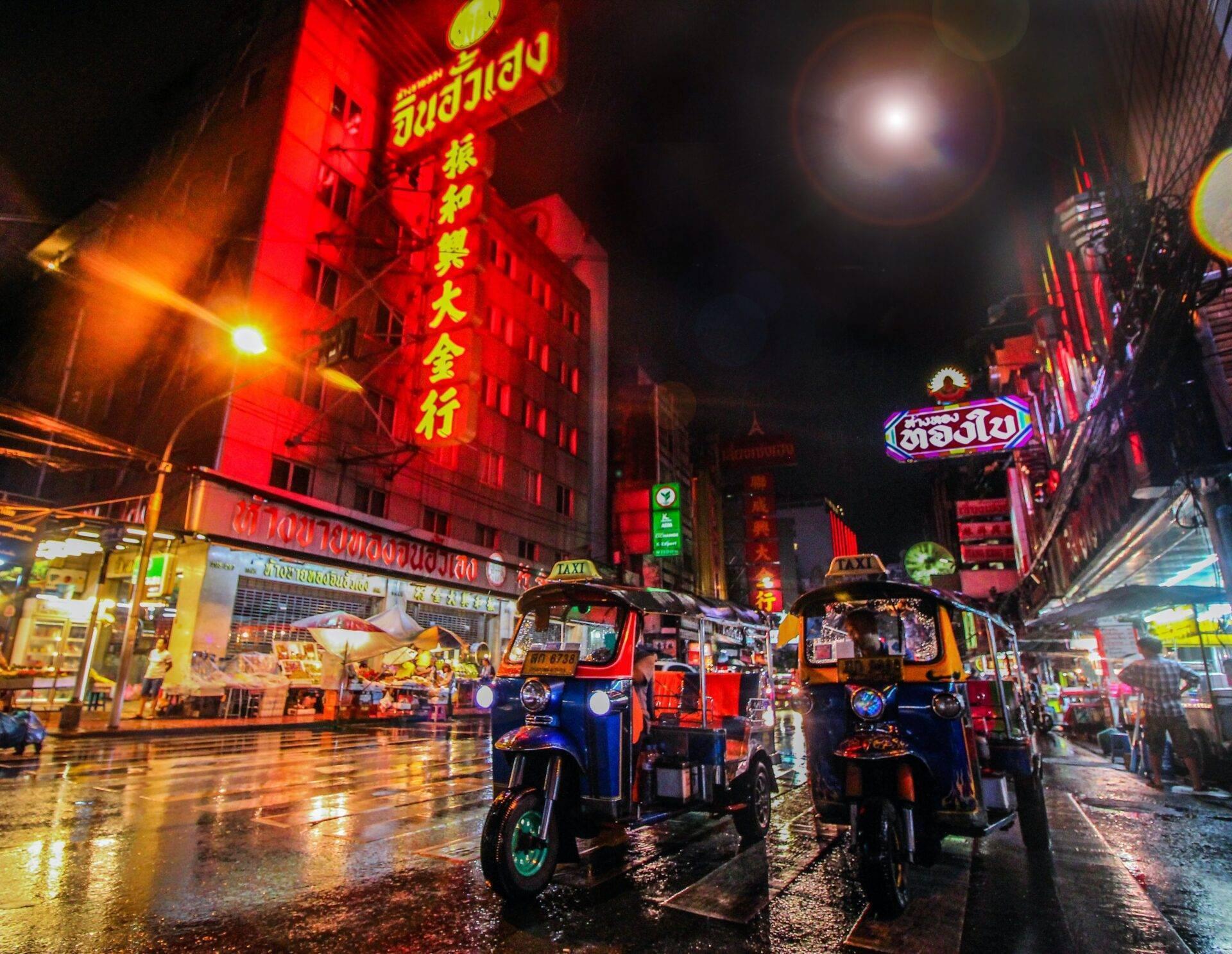 Calle animada de noche en Tailandia con tuk-tuks azules, carteles luminosos rojos y amarillos, y suelo mojado que refleja las luces