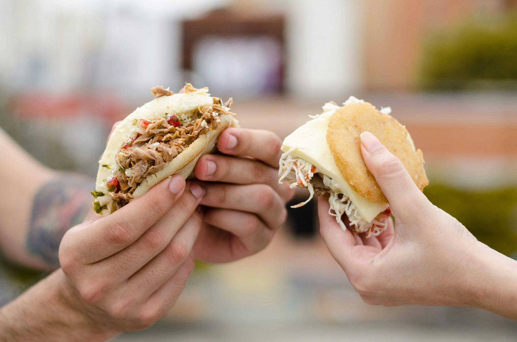 Dos manos sosteniendo bocadillos rellenos de carne desmechada en una calle empedrada de fondo difuminado