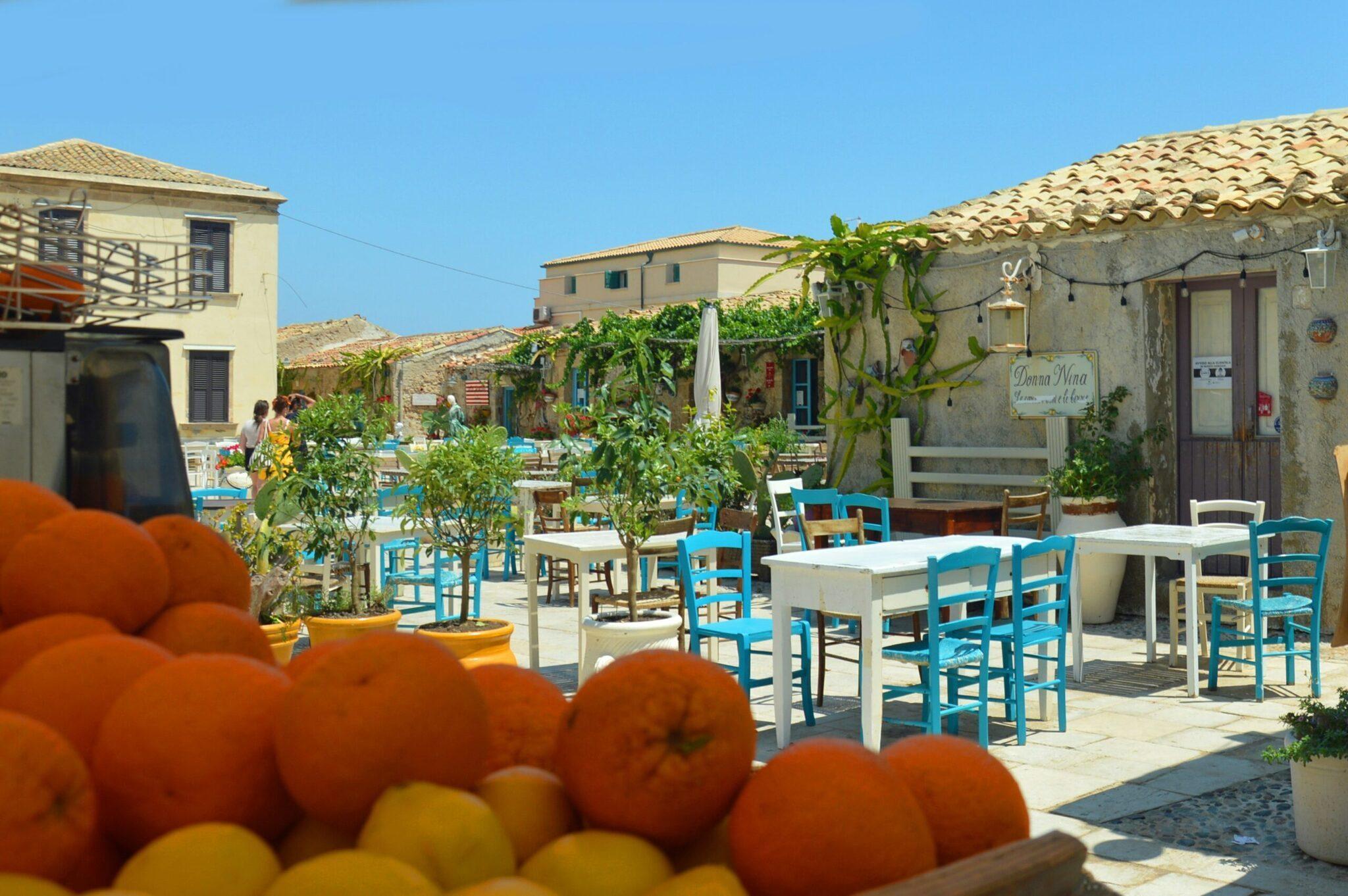 Una terraza mediterránea con mesas azules y blancas, cítricos frescos en primer plano y un pueblo siciliano típico al fondo