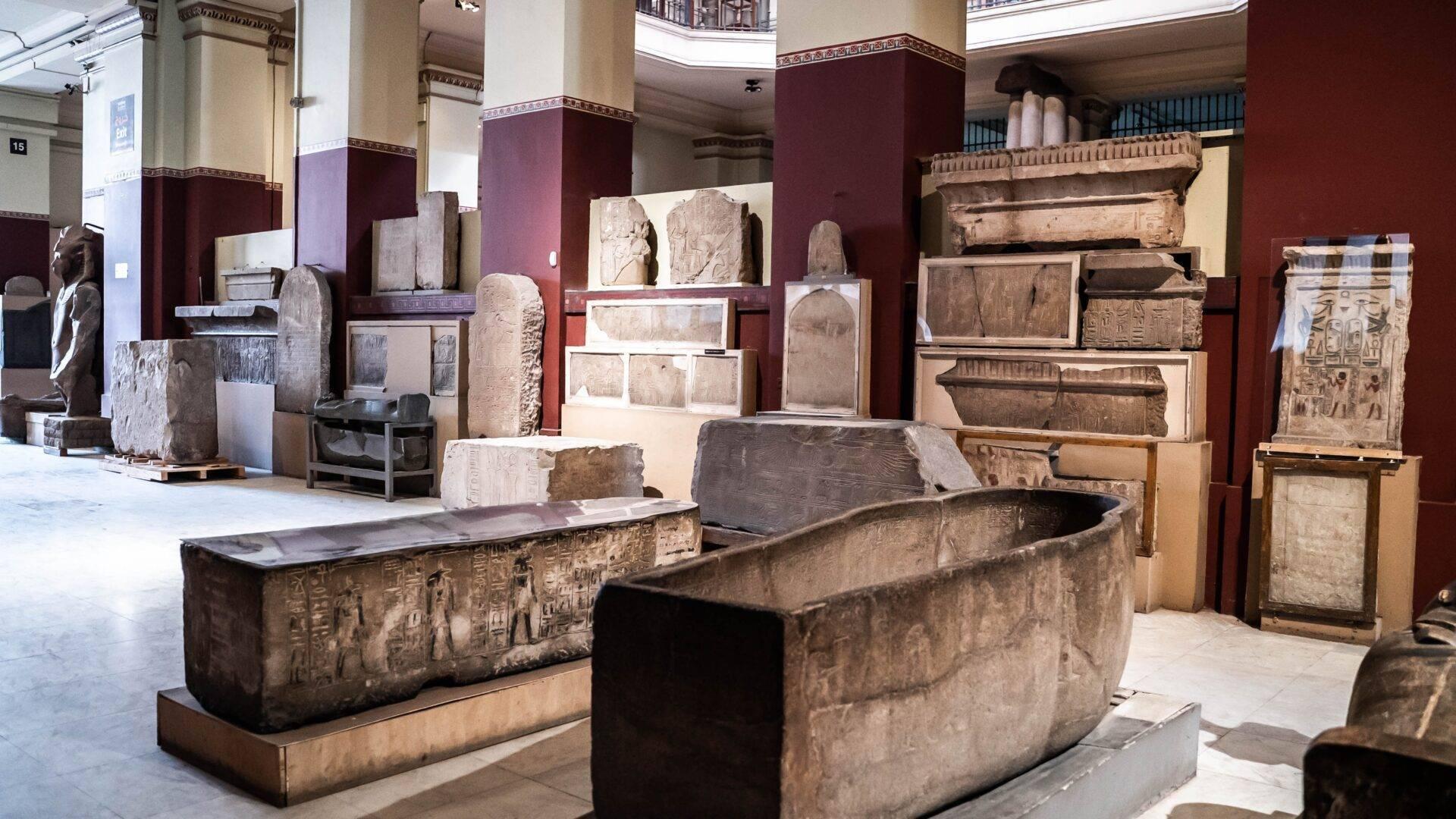 Sarcofagi egizi antichi e bassorilievi esposti in una galleria di museo dalle pareti bordeaux