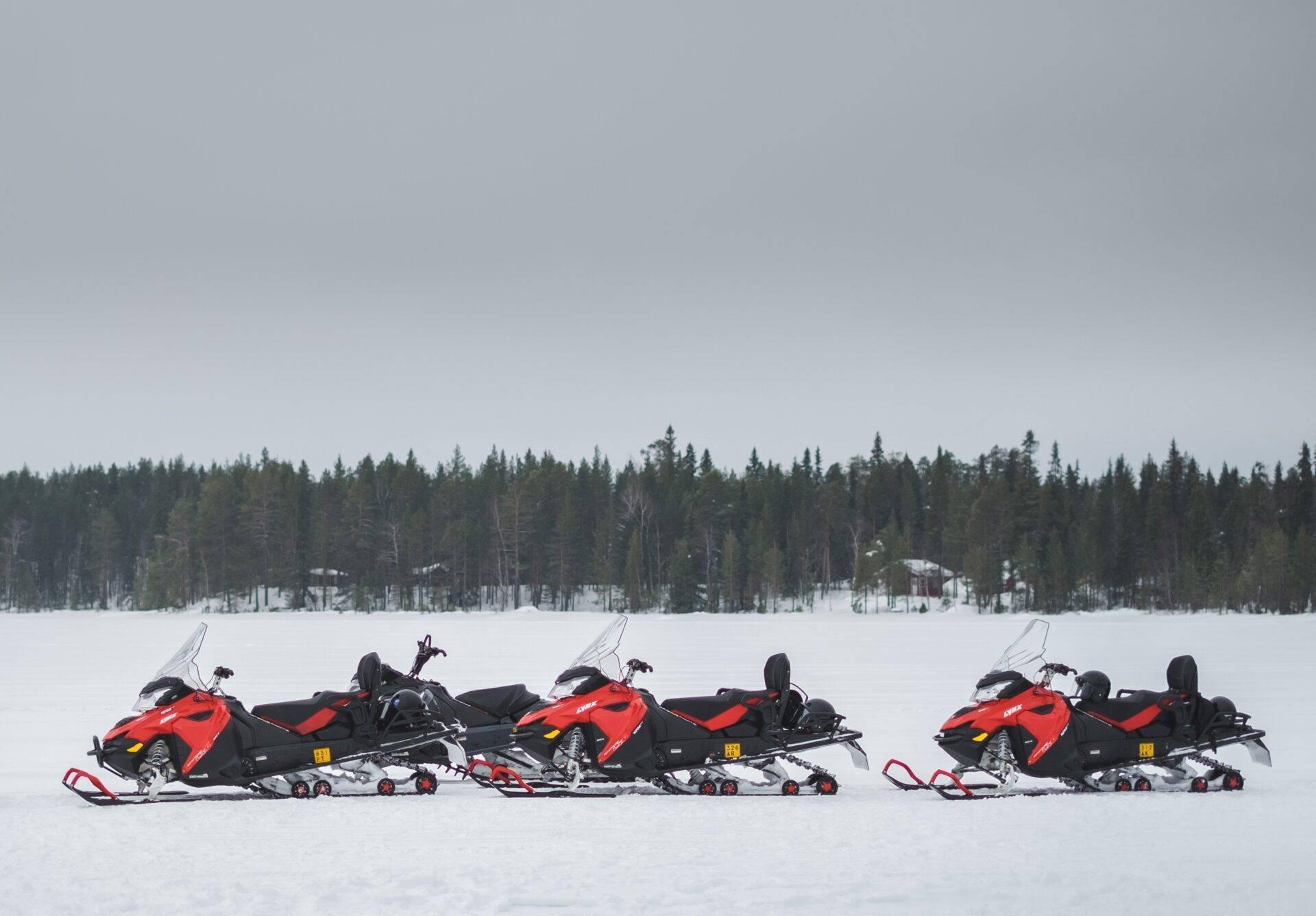 Tres motos de nieve rojas alineadas sobre un lago helado frente a un bosque de abetos nevados