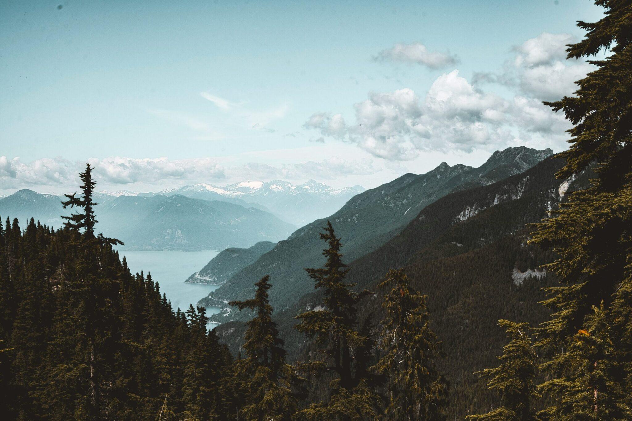 Panoramic view of an alpine valley with a mountain lake, conifer forests and snow-dusted peaks in the background