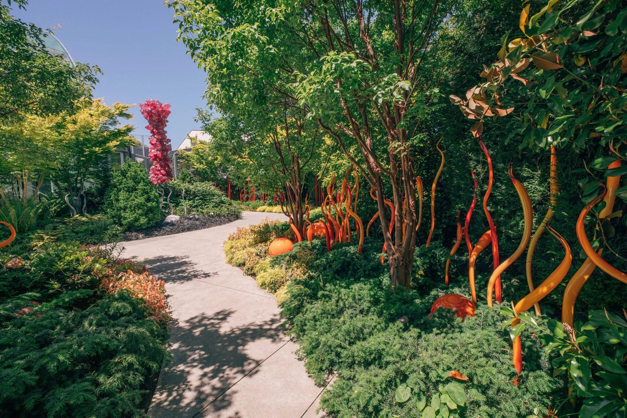 Paseo de jardín moderno flanqueado por árboles con troncos pintados en rojo y naranja vivo, con esculturas de colores