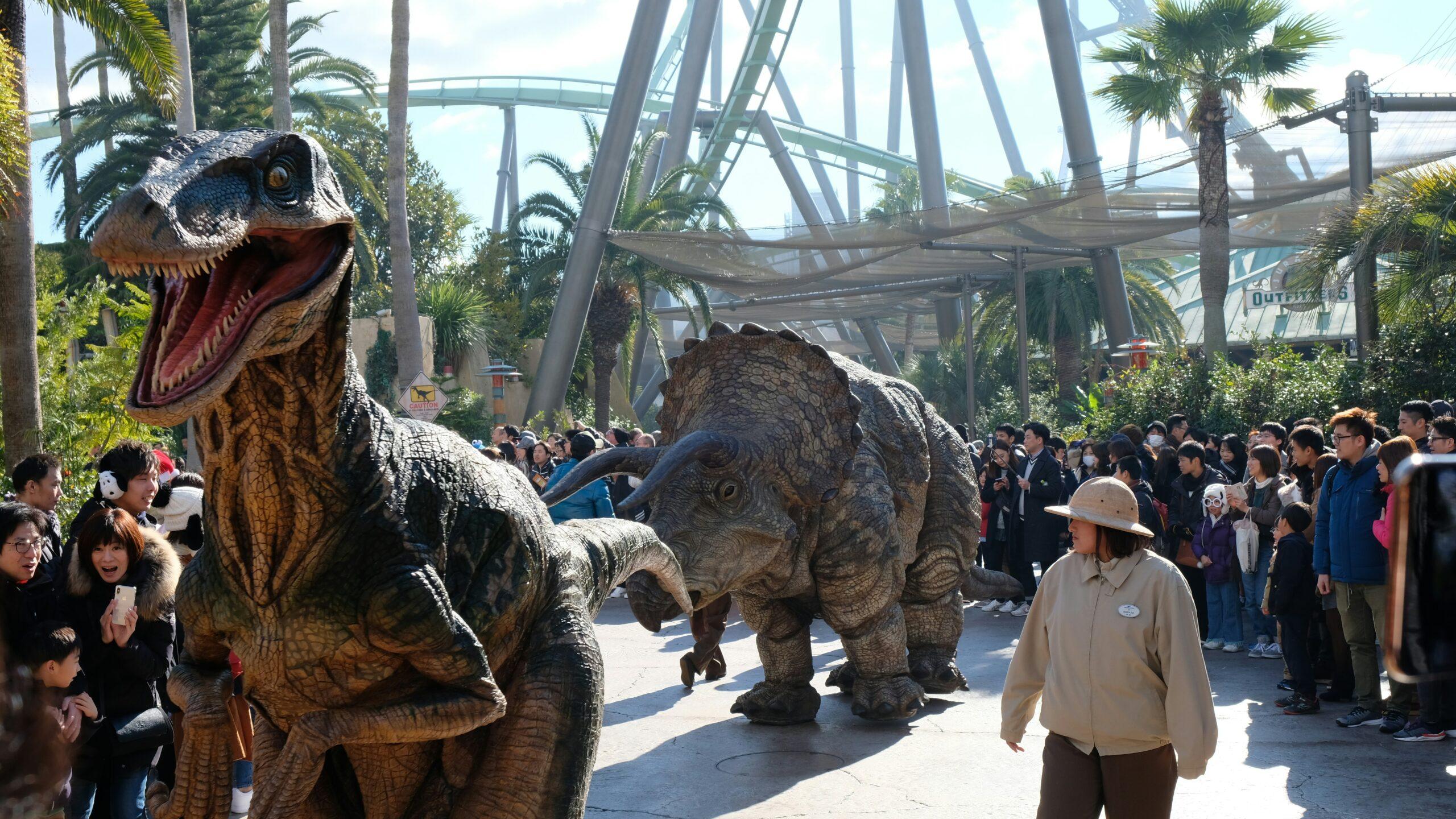 Sfilata di dinosauri animatronici in un parco a tema con folla di visitatori e strutture metalliche di montagne russe sullo sfondo