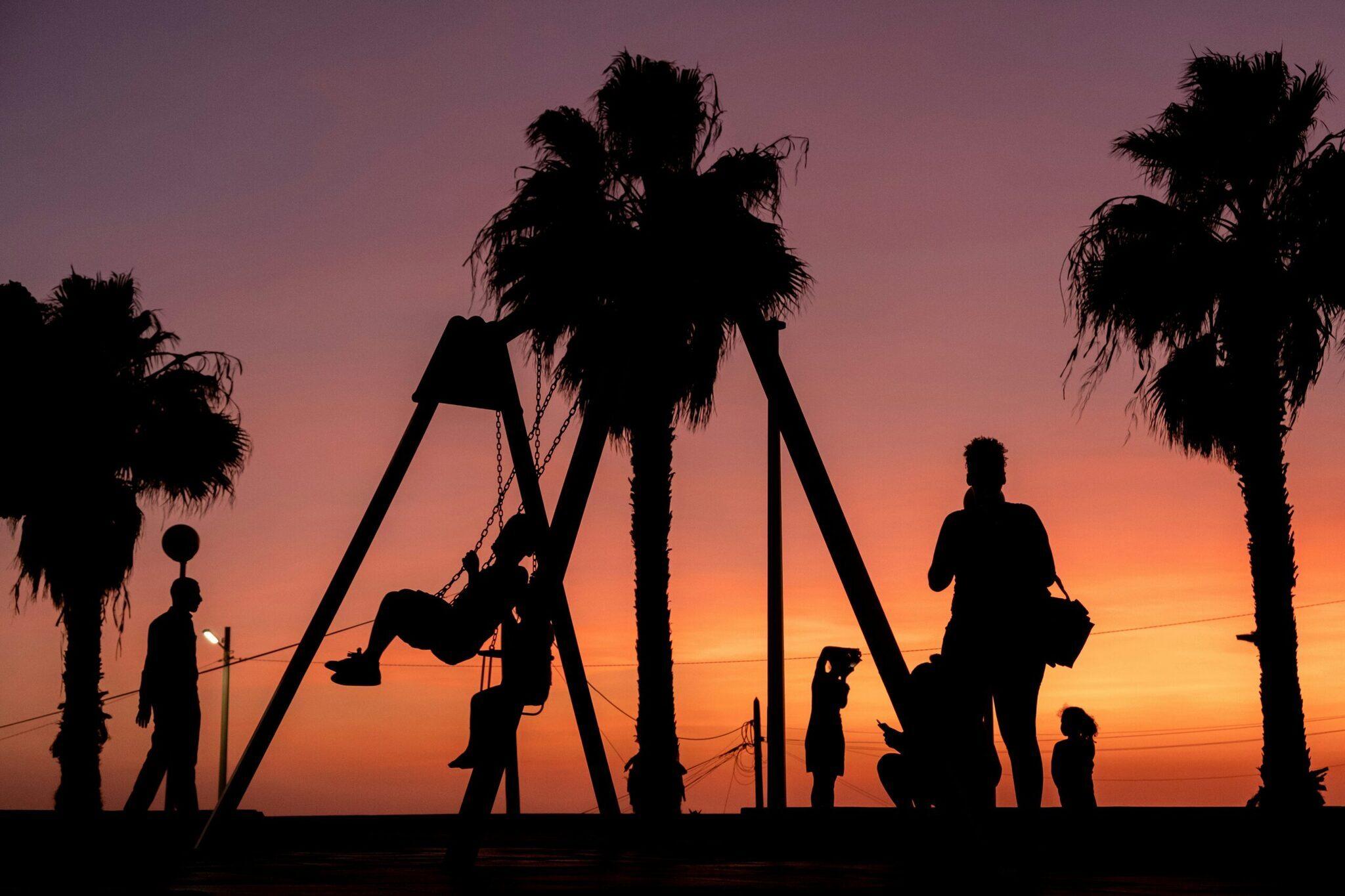 Siluetas de niños jugando en un columpio al atardecer con palmeras y cielo naranja