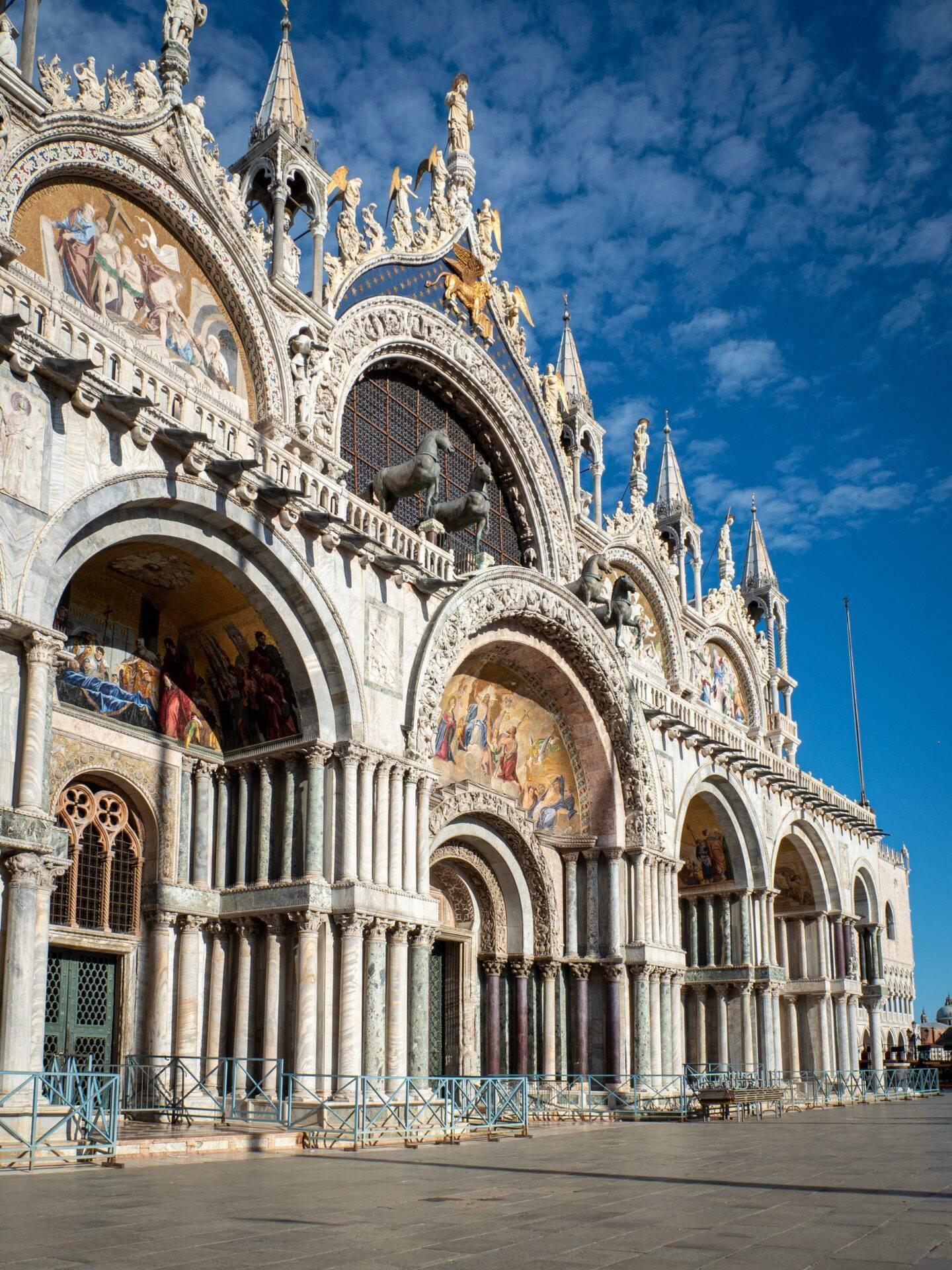 Facciata ornata di una basilica veneziana con arcate gotiche, sculture dorate e mosaici su sfondo di cielo azzurro