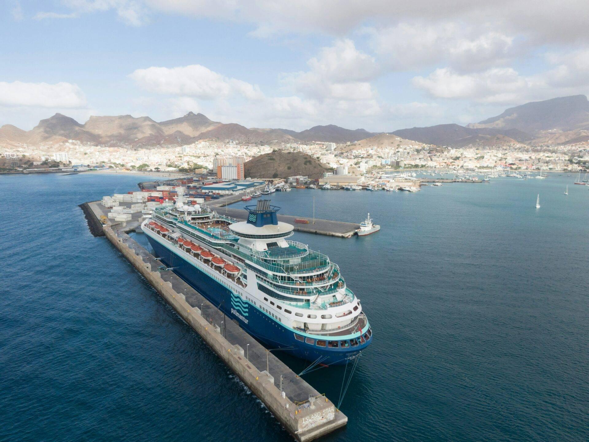 A large cruise ship docked in the port of a coastal town with arid mountains in the background