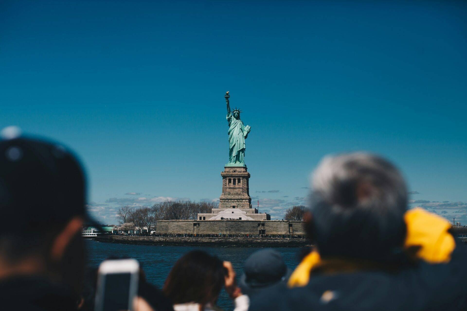 Turisti fotografano l’iconica Statua della Libertà sulla sua isola, sotto un cielo azzurro limpido