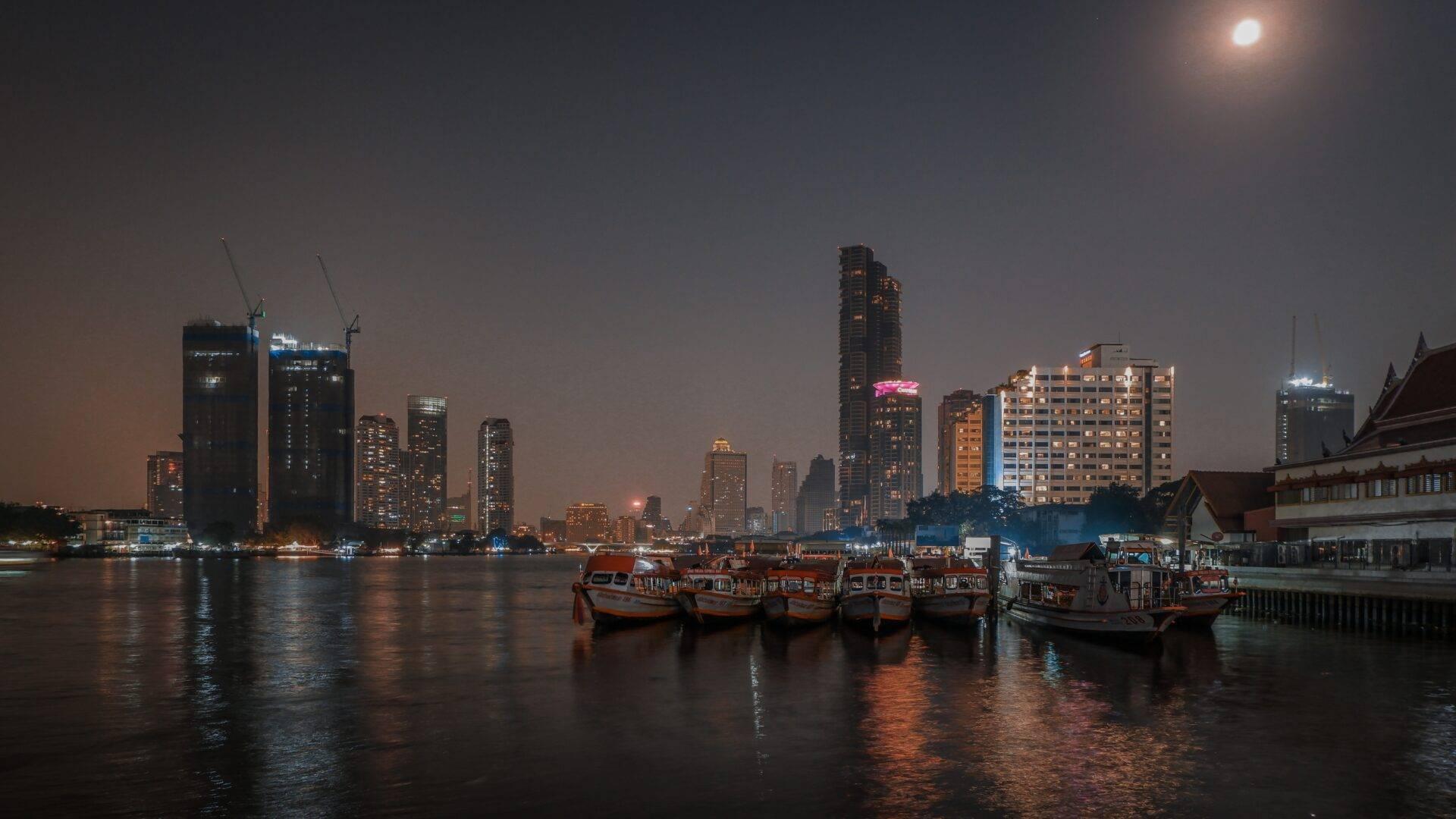 Skyline urbano illuminato di notte con battelli tradizionali rossi ormeggiati davanti a un’architettura moderna sul fiume