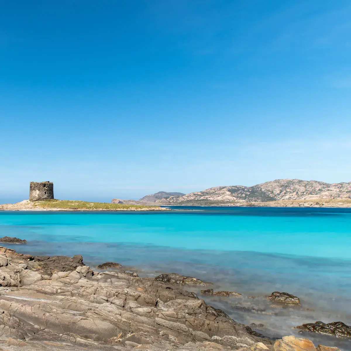 Ein alter Steinturm auf einer felsigen Küste vor türkisfarbenem Meer und kargen Hügeln