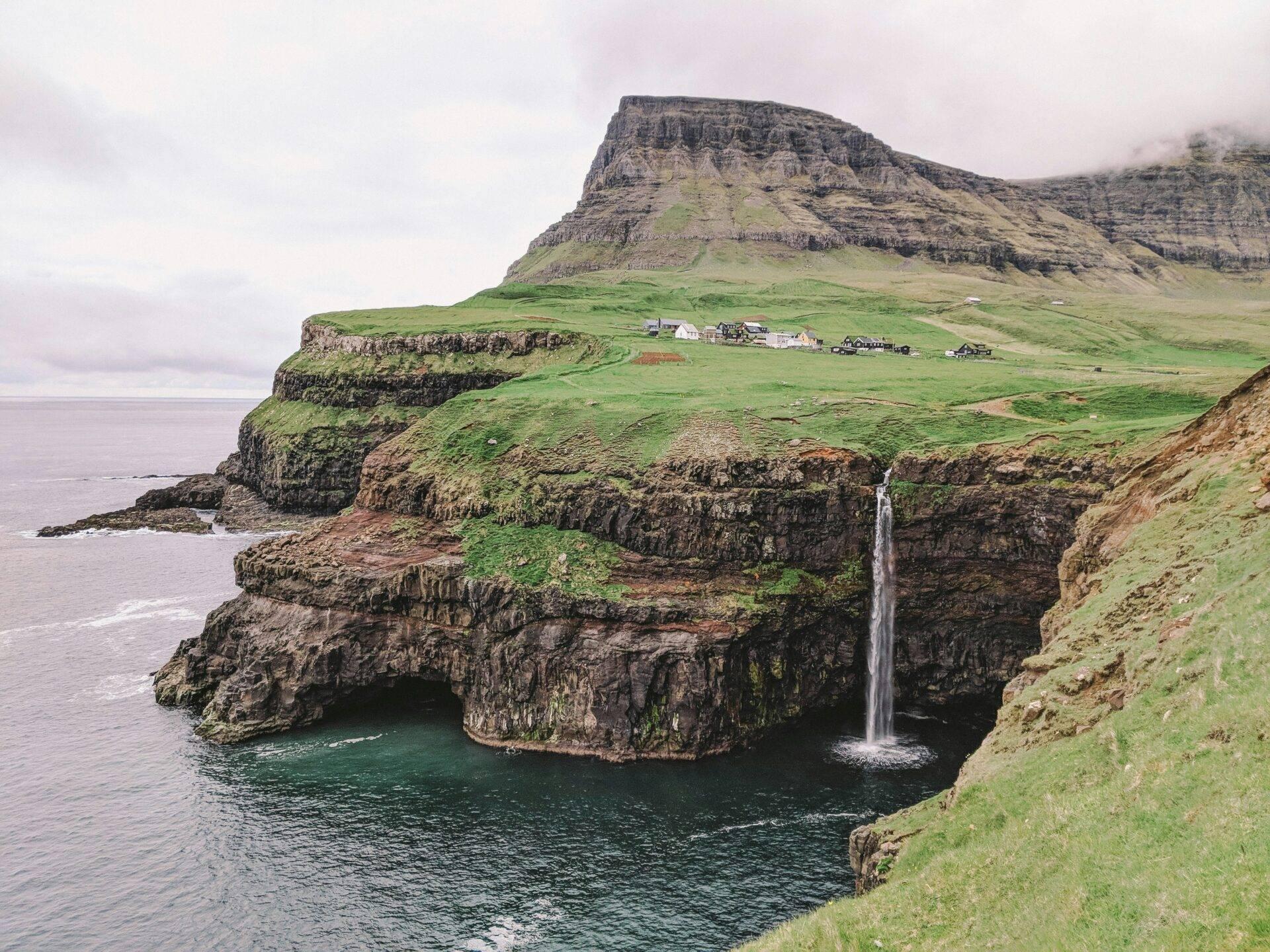 Bergige Küstenlandschaft mit Wasserfall, grünen Klippen und vereinzelten Höfen unter bewölktem Himmel