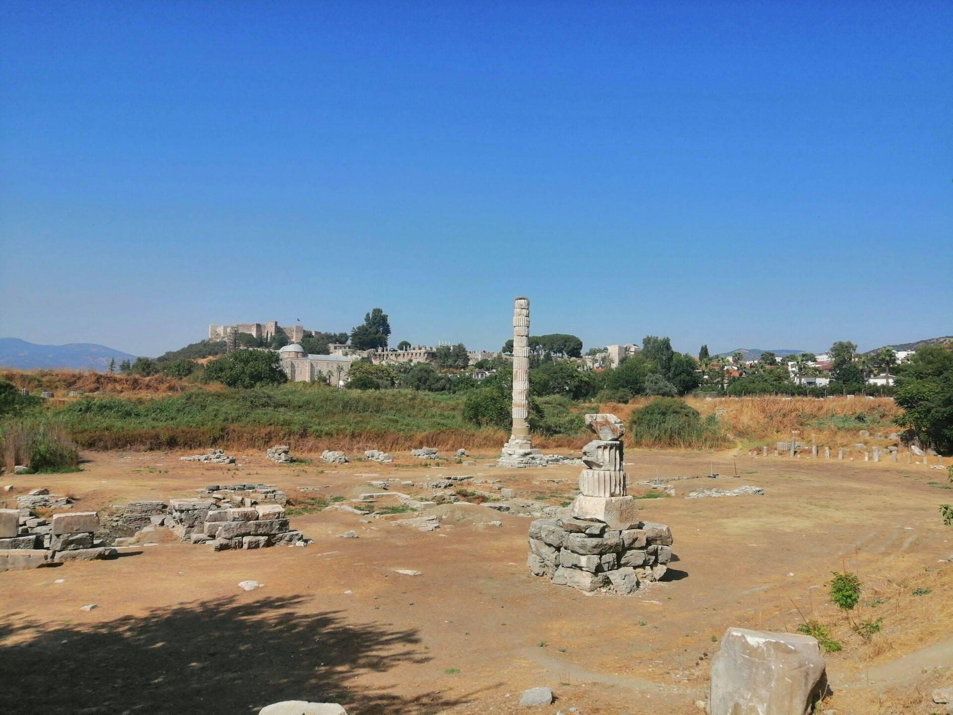 Rovine antiche con colonna isolata, tempio sullo sfondo e montagne sotto un cielo sereno