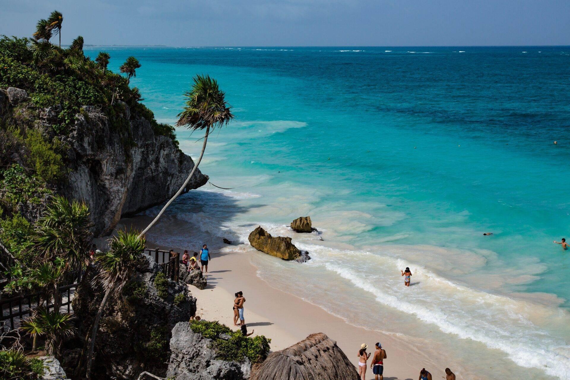 Weißer Sandstrand mit Felsen, Kalksteinklippe und Palme, Badegäste im kristallklaren türkisfarbenen Wasser