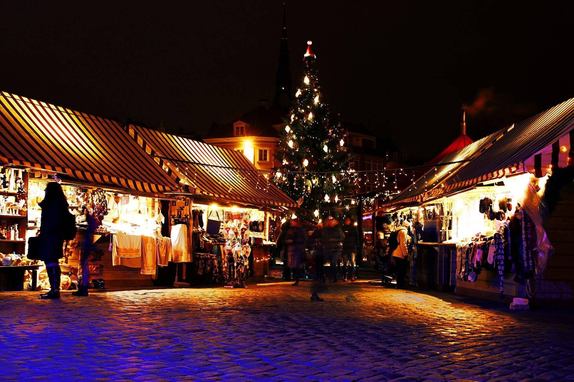 Un mercado navideño iluminado de noche con abeto decorado, casetas y visitantes paseando