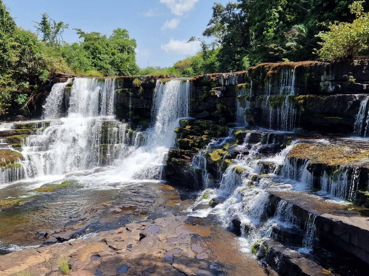 Una spettacolare cascata a più livelli con rocce muschiate, circondata da vegetazione lussureggiante e un cielo sereno