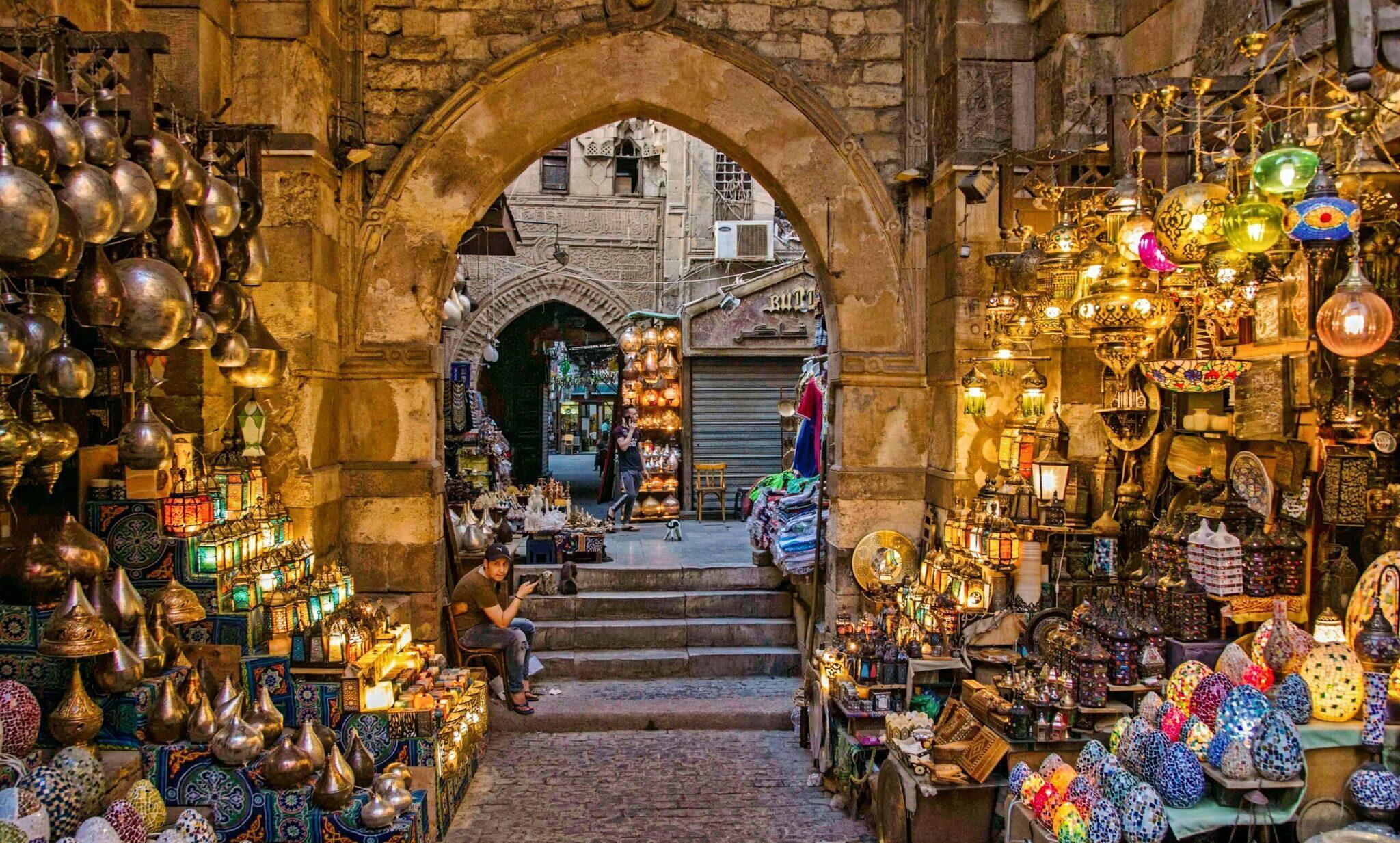 Callejón cubierto de un mercado oriental iluminado, flanqueado por faroles dorados y artesanía de colores, con un arco de piedra
