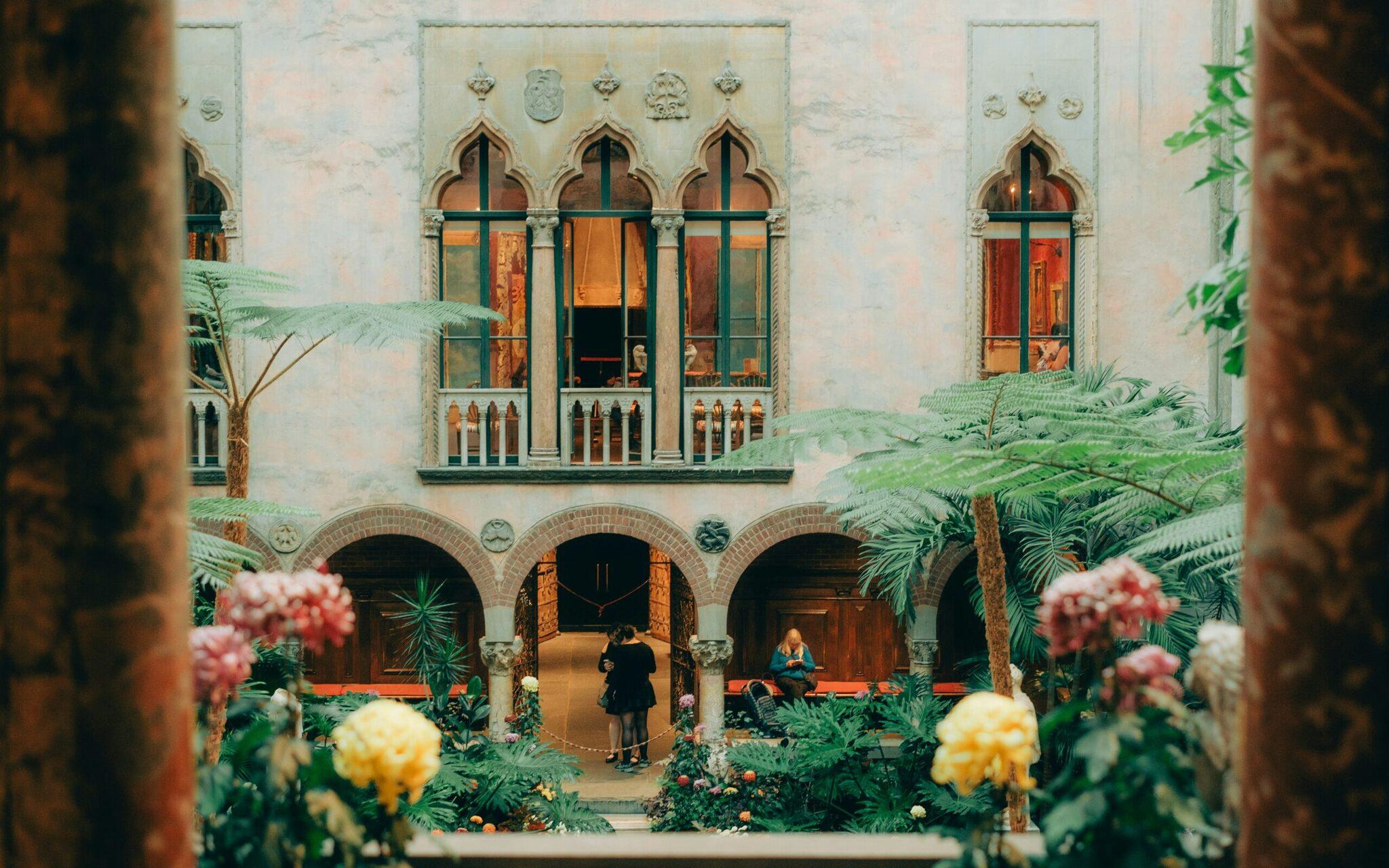 An interior courtyard with brick arches, Gothic windows, tropical plants and visitors strolling in a Mediterranean atmosphere