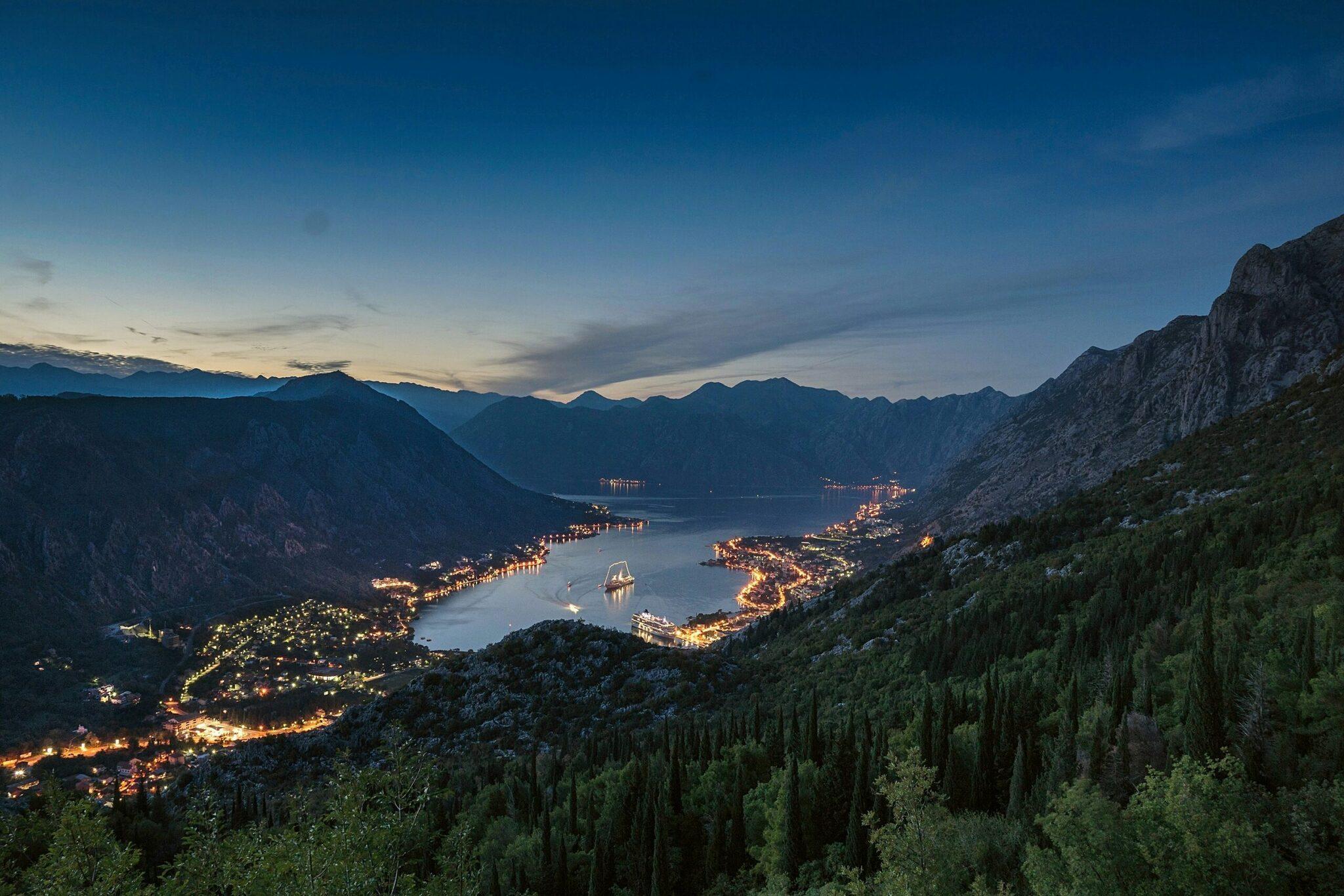 Vista notturna di una baia costiera incorniciata da montagne con città illuminate e una barca a vela al crepuscolo