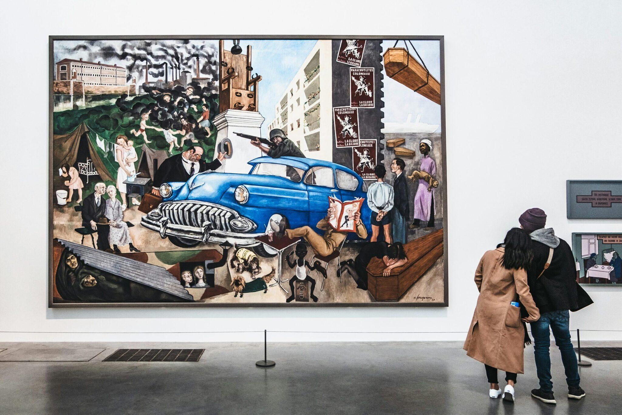 Dos visitantes observan un mural colorido que representa un coche azul vintage y escenas urbanas en un museo
