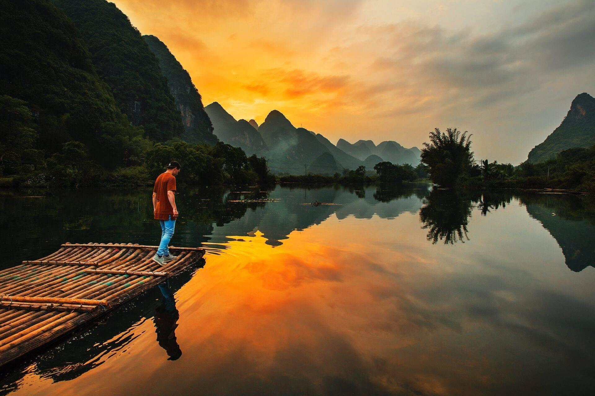 Un uomo in camicia arancione su una zattera di bambù contempla un fiume tranquillo circondato da montagne carsiche al tramonto