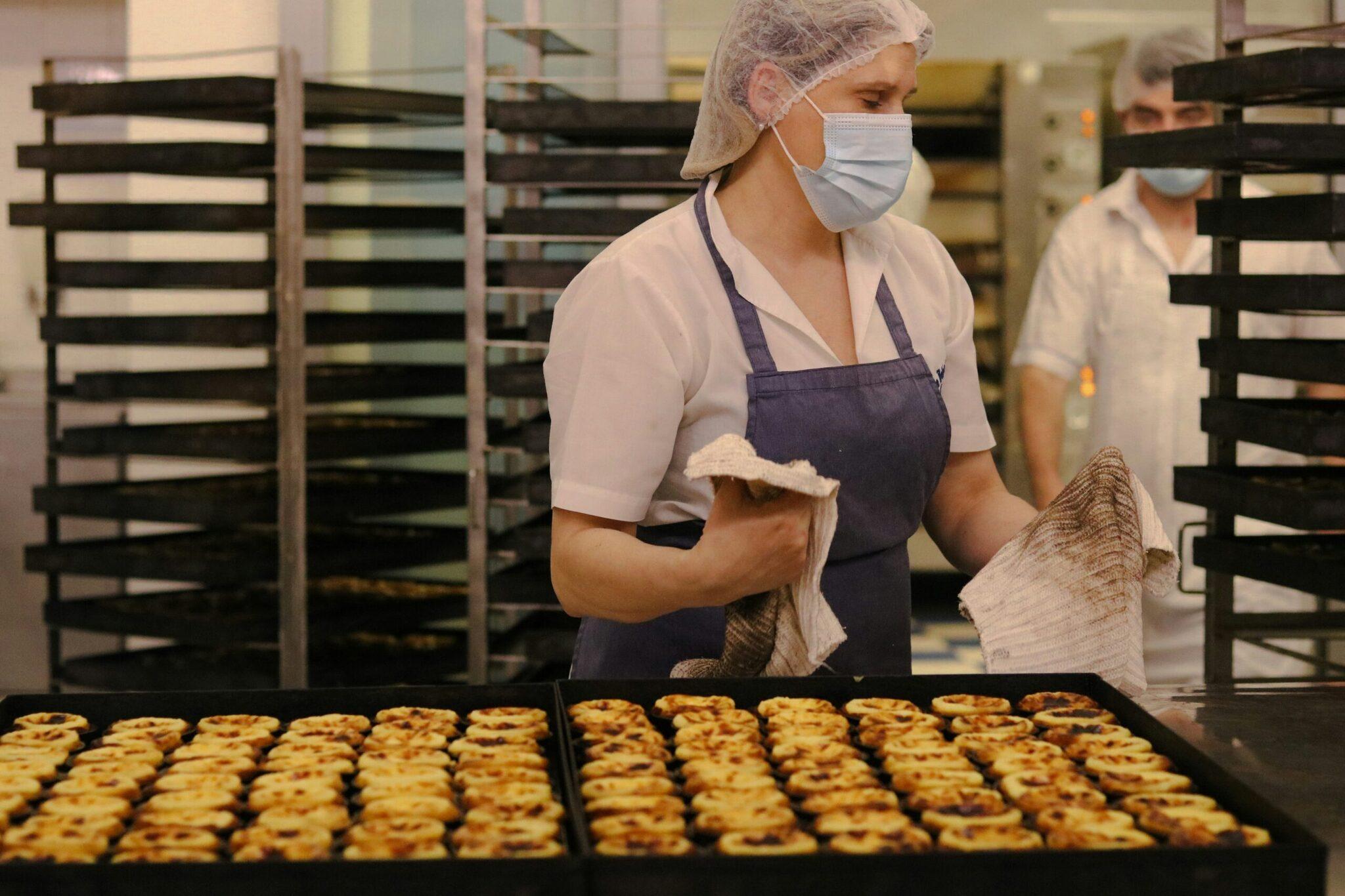 Eine Bäckereimitarbeiterin in Arbeitskleidung präsentiert frisch gebackene Cremetörtchen in einer Konditorei