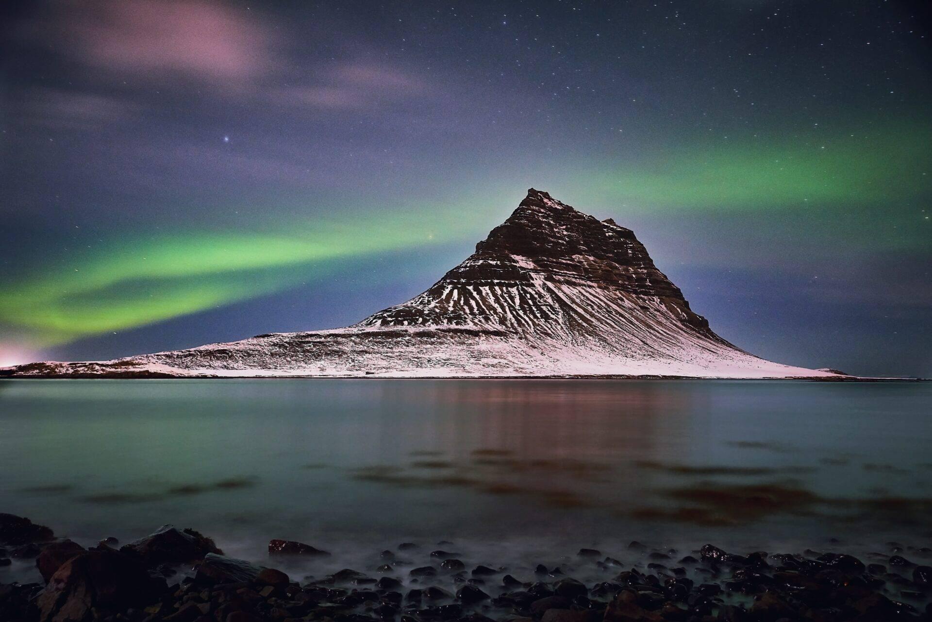 Una montaña nevada se refleja en una bahía helada bajo una aurora boreal verde y un cielo estrellado