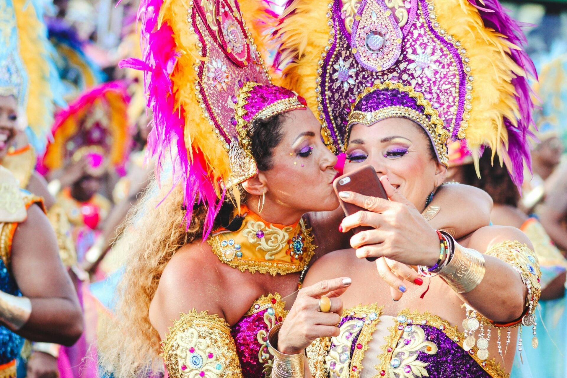 Due ballerine in costumi colorati e piumati partecipano a un carnevale tropicale, una scatta un selfie