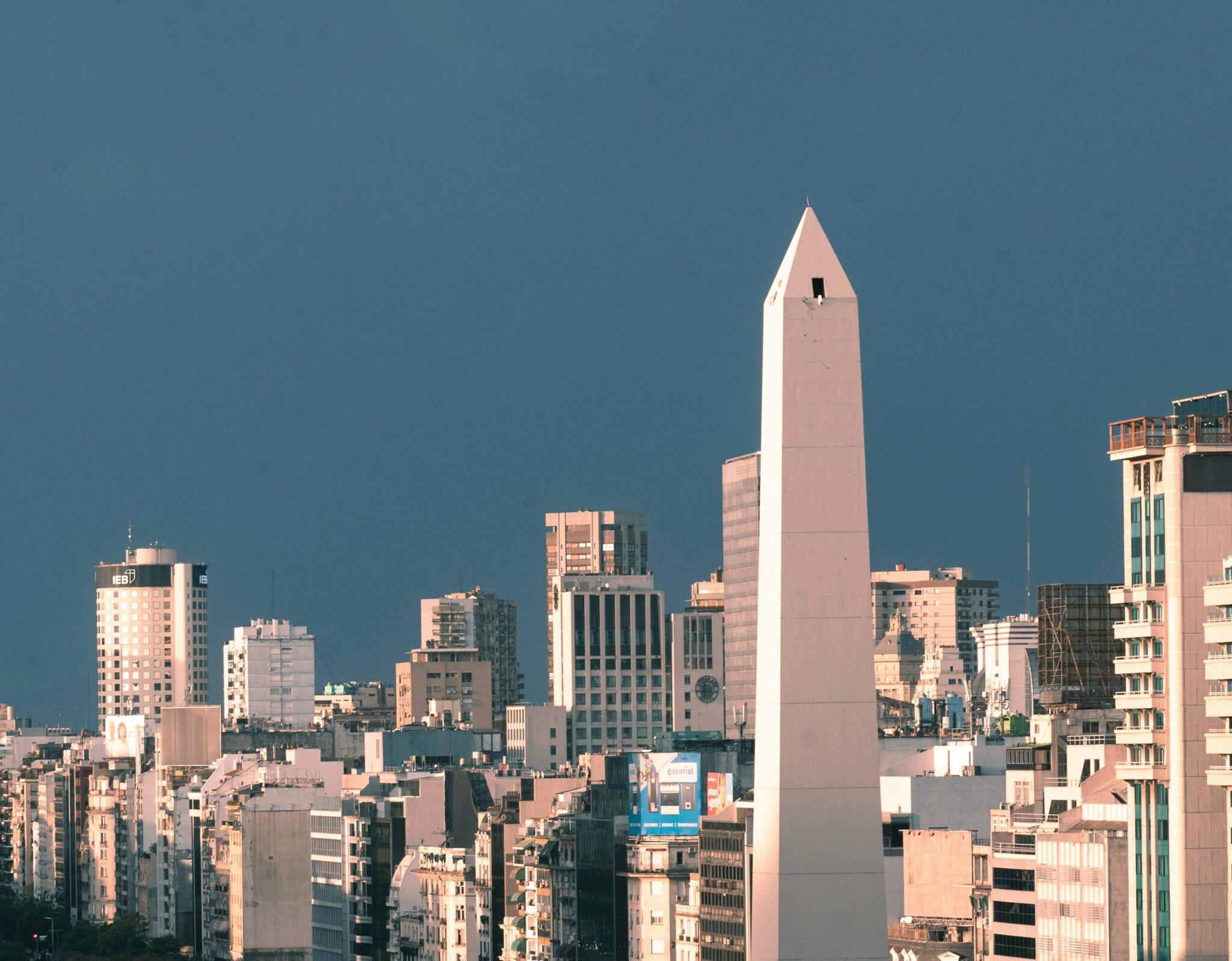 Skyline urbano con un obelisco bianco che svetta sugli edifici sotto un cielo azzurro e terso