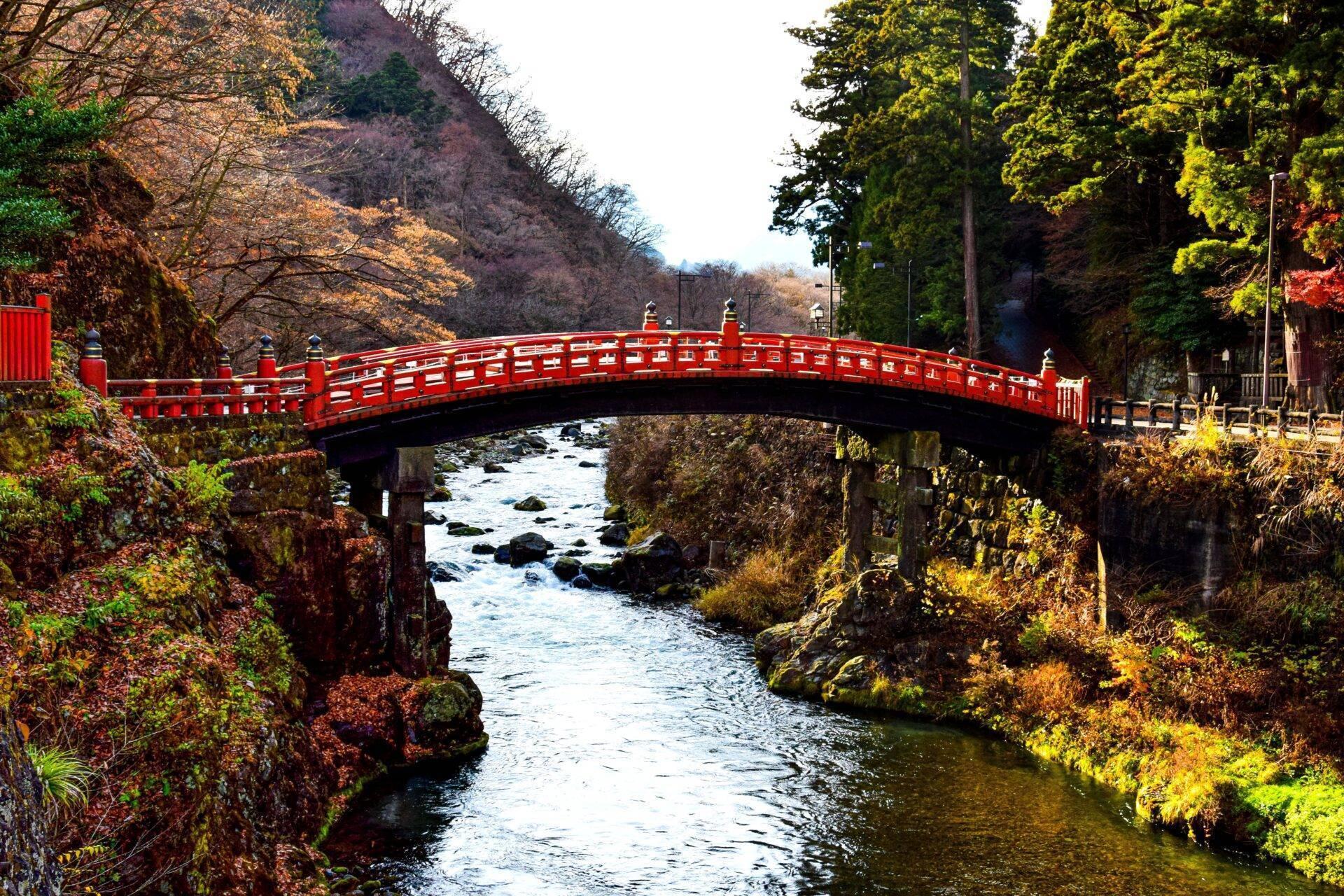 Eine rote Bogenbrücke überspannt einen felsigen Fluss in einer herbstlich bewaldeten japanischen Landschaft
