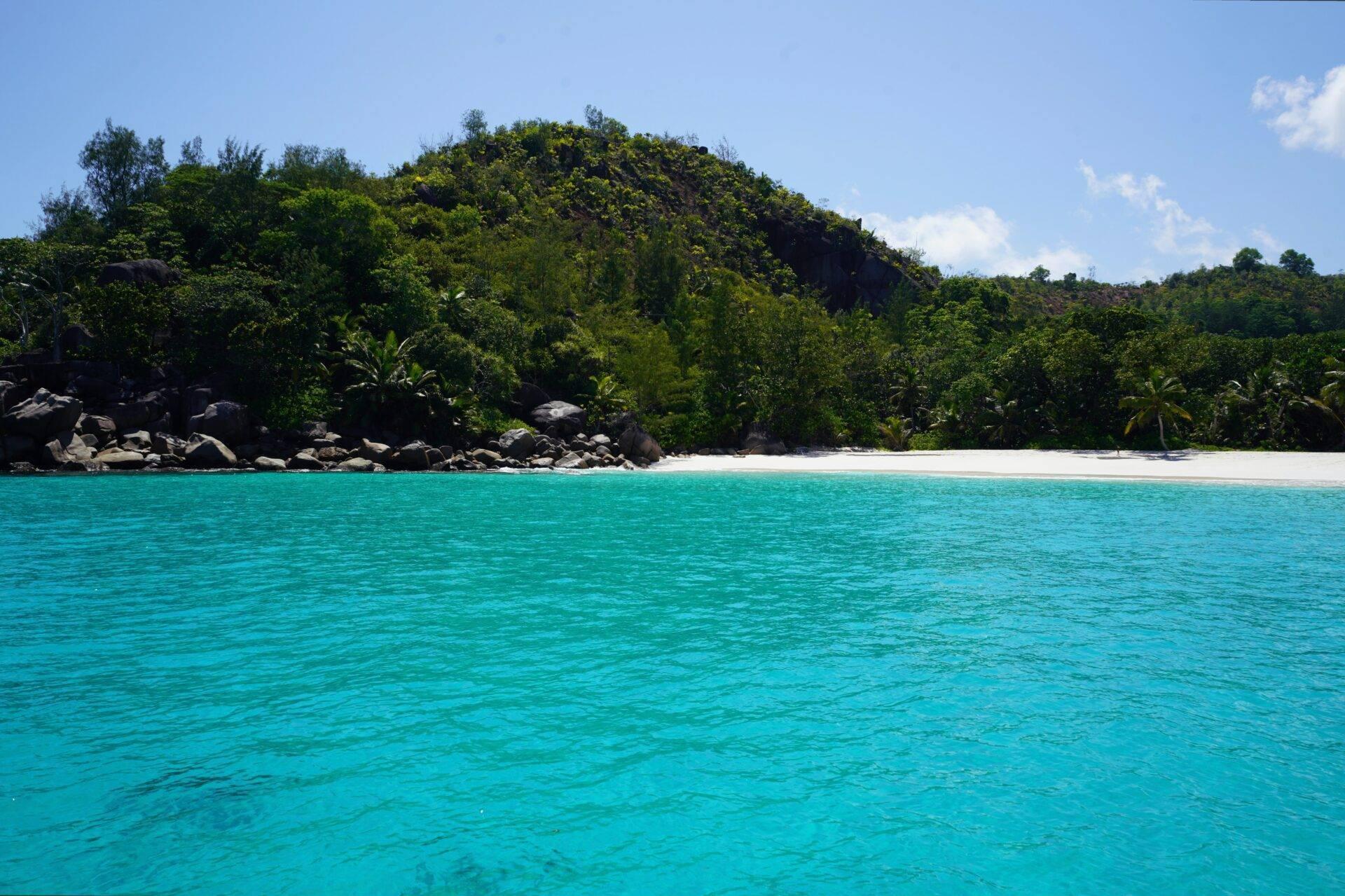 Ein weißer Strand mit türkisfarbenem Wasser, gesäumt von Felsen und üppiger Tropenvegetation unter blauem Himmel