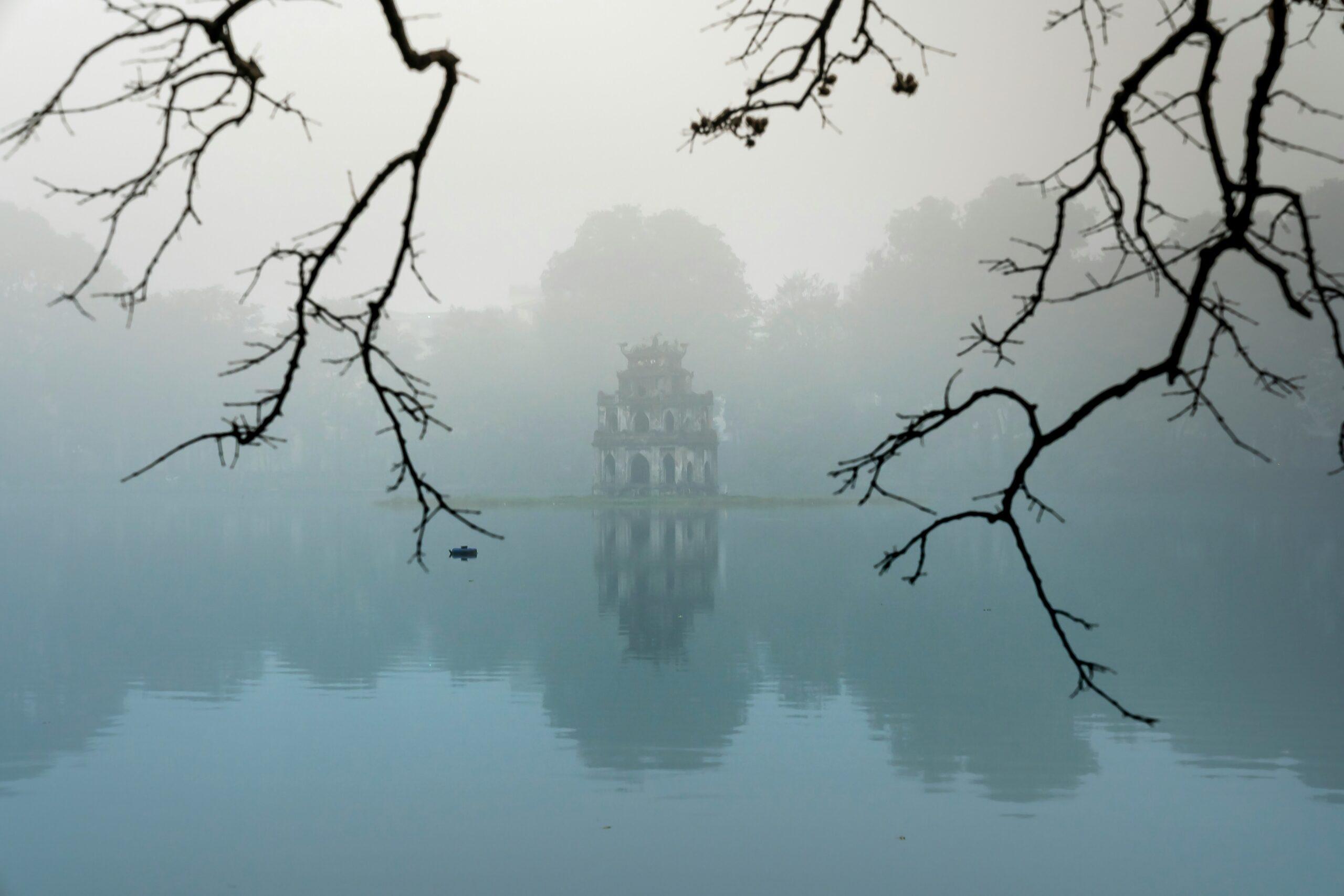 Una torre histórica se refleja en un lago tranquilo bajo una densa niebla, enmarcada por ramas desnudas
