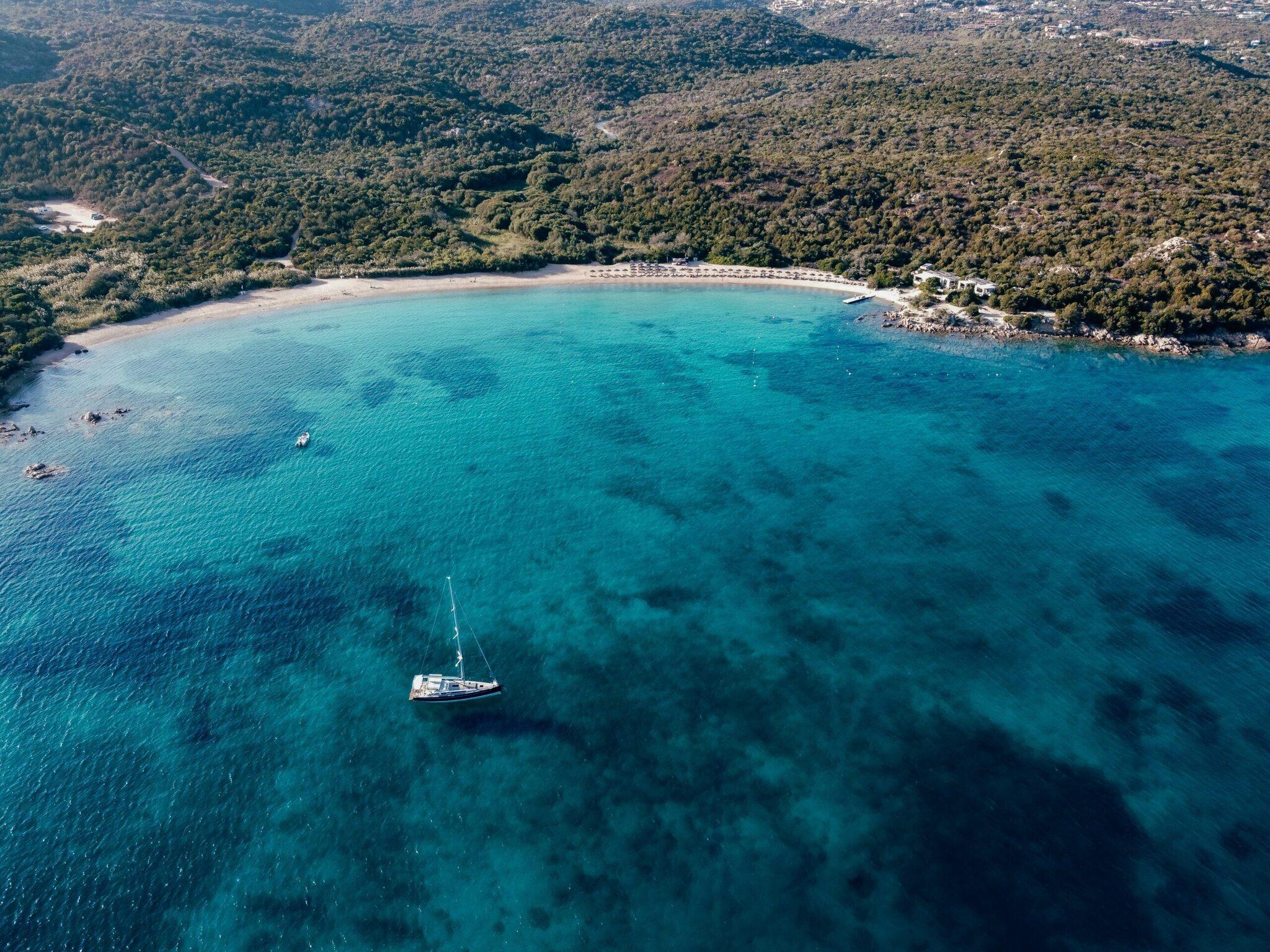 Vista aérea de una bahía mediterránea de aguas turquesas con velero, playa de arena y bosques costeros