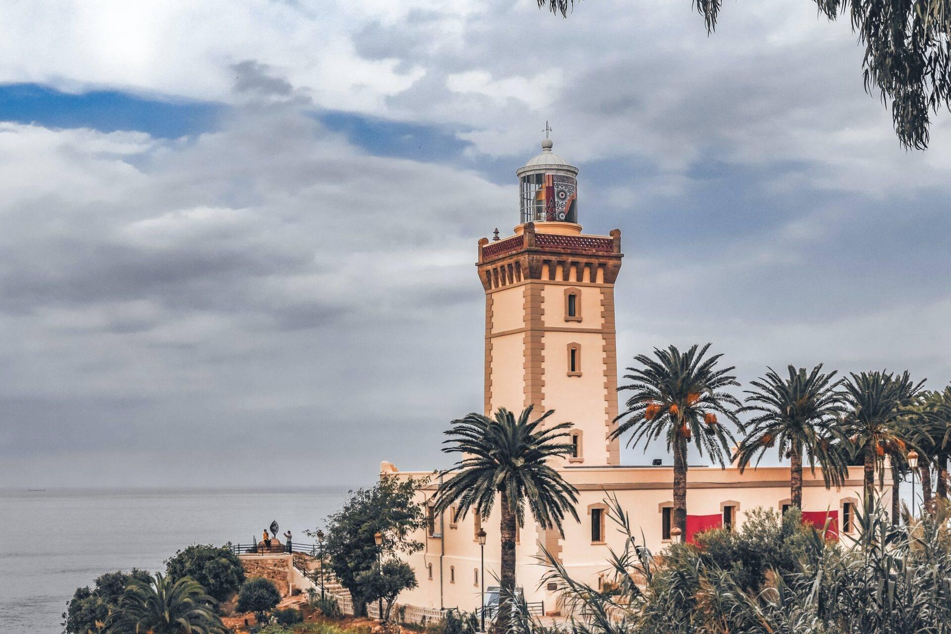 Weißer Küstenleuchtturm mit Laternenturm, umgeben von Palmen mit Blick auf das Mittelmeer unter bewölktem Himmel