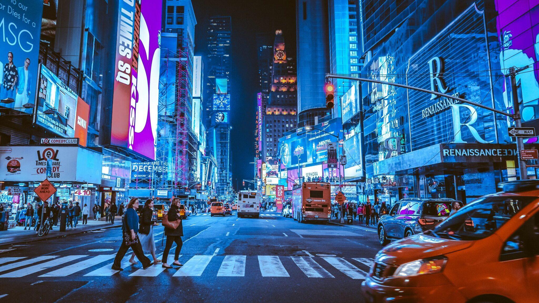 Una calle urbana animada iluminada con publicidad de colores, peatones cruzando y taxis naranjas de noche