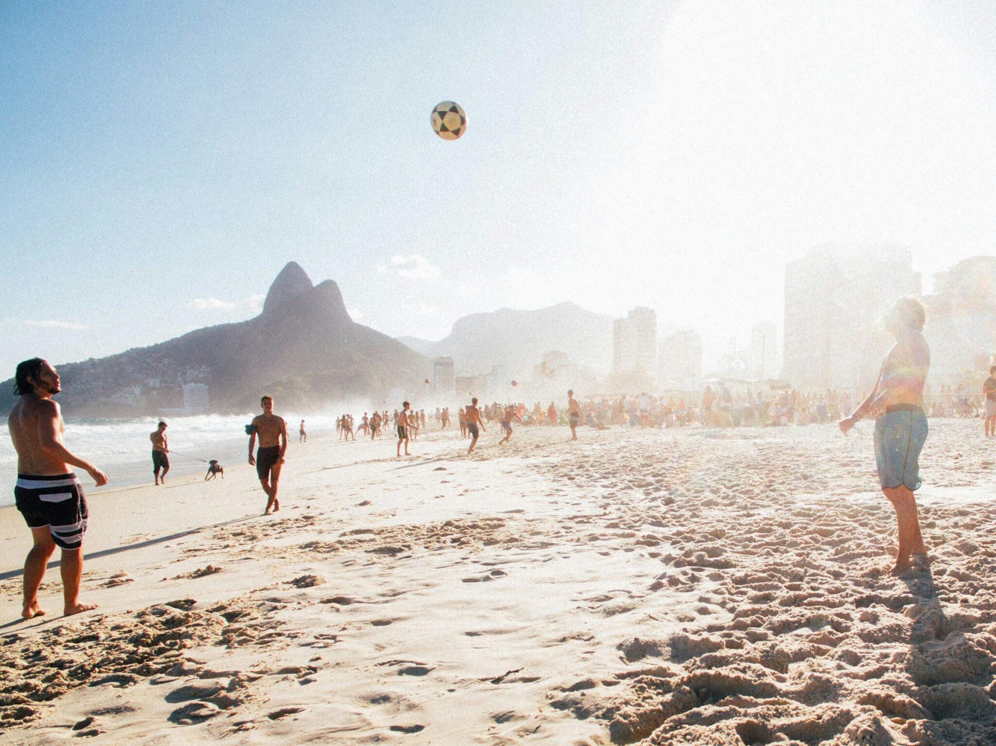 Grupo de personas jugando con el balón en una playa de arena fina con la montaña característica y edificios al fondo