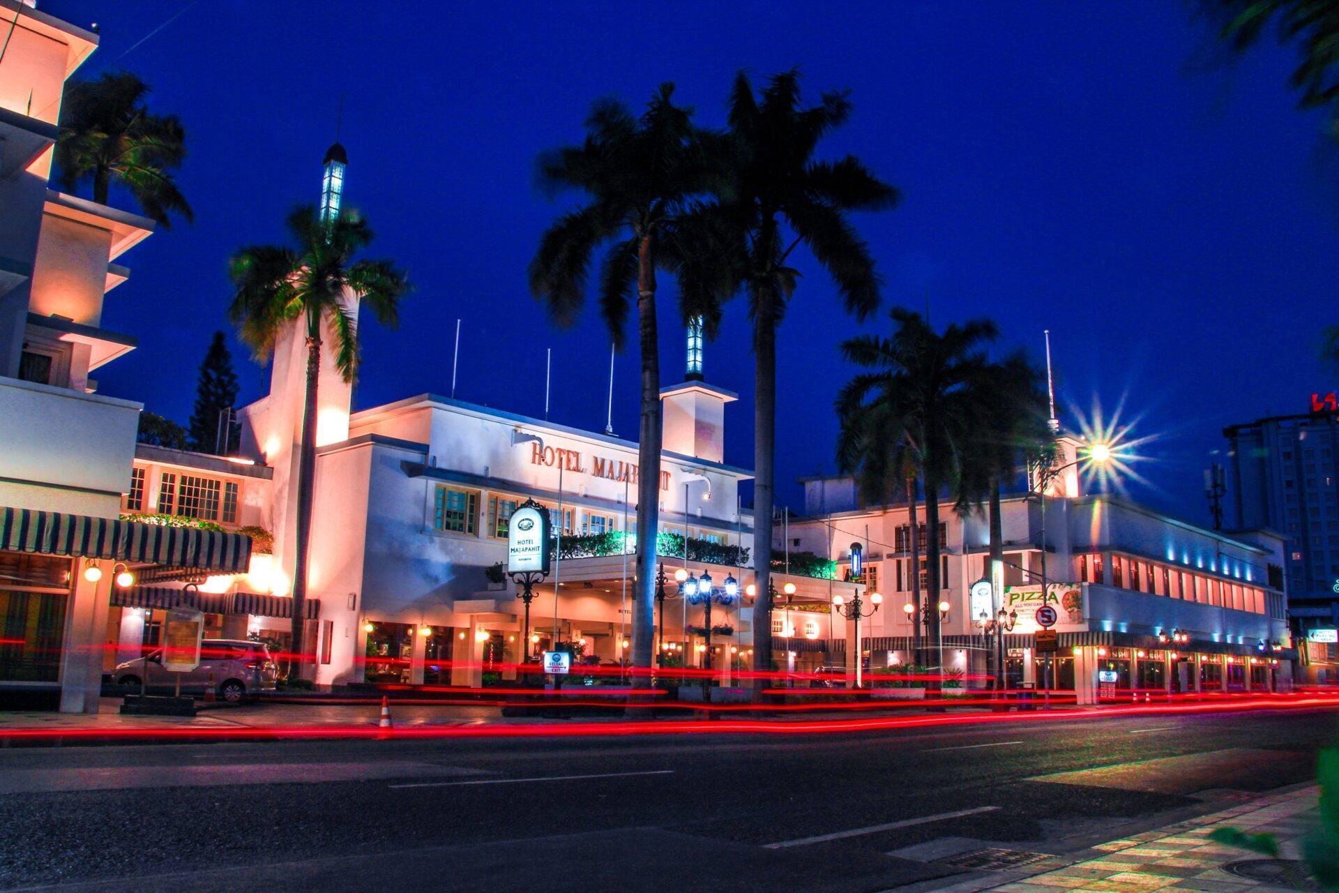Facciata illuminata di un hotel coloniale di notte con palme, architettura art déco e scie rosse dei fari
