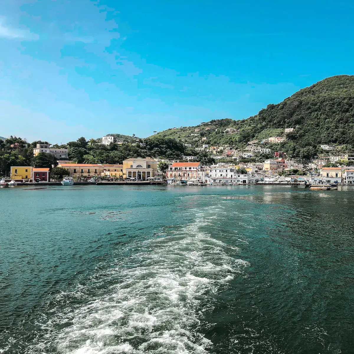Blick vom Meer auf ein buntes mediterranes Küstendorf mit Pastellhäusern und bewaldeten Hügeln