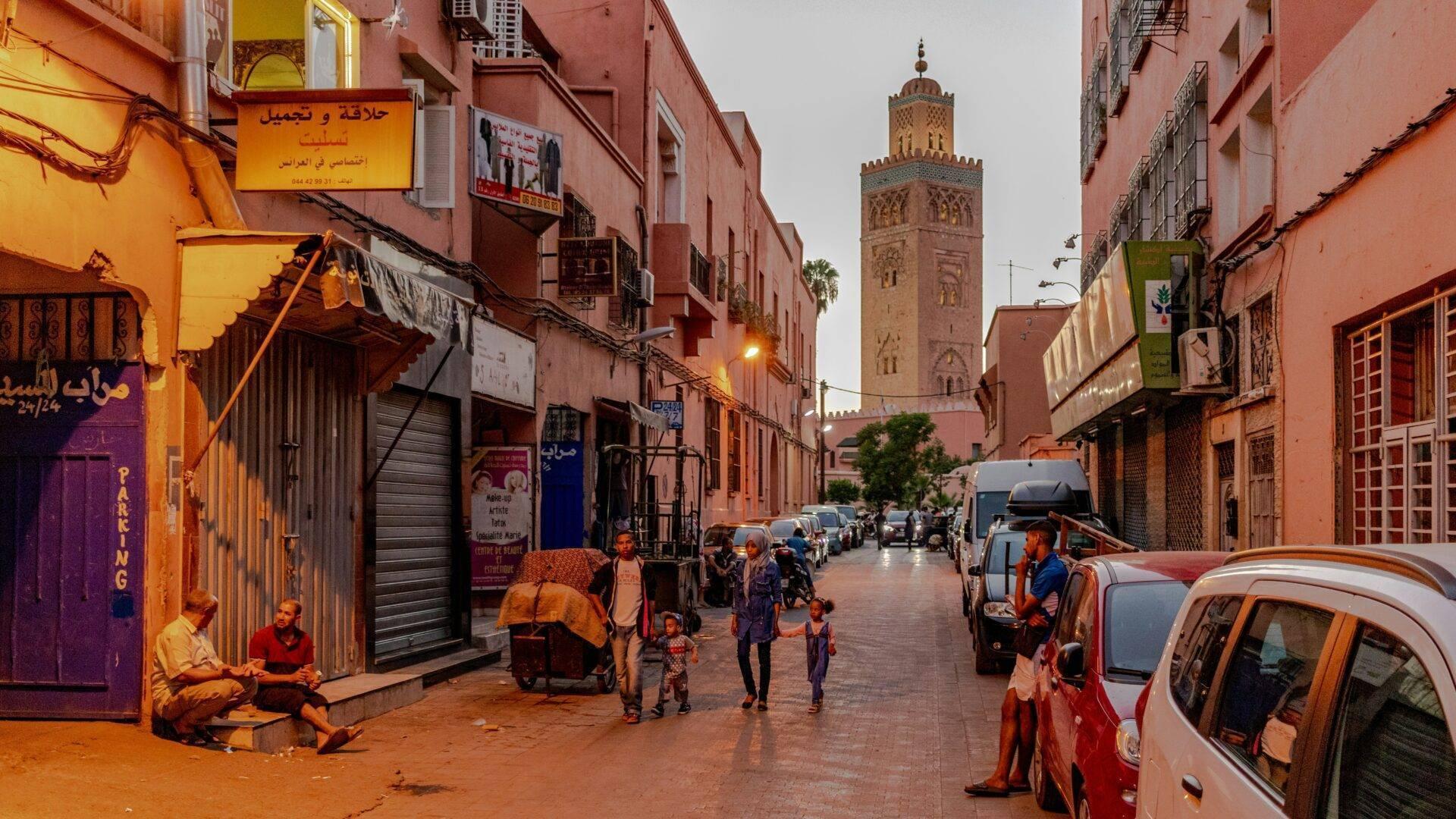 Una calle comercial animada de una medina marroquí con una mezquita al fondo, peatones y coches