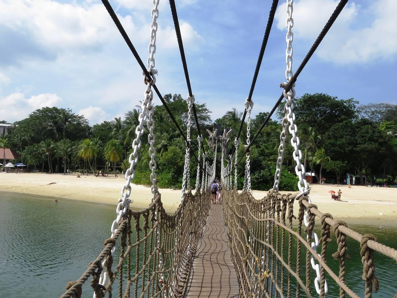 Eine Hängebrücke aus Seilen und Metallketten über einem tropischen Strand mit üppiger Vegetation
