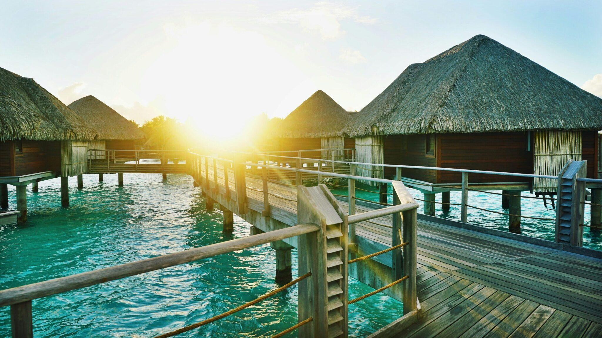 Überwasserbungalows mit Strohdächern über einer türkisfarbenen Lagune im Abendlicht
