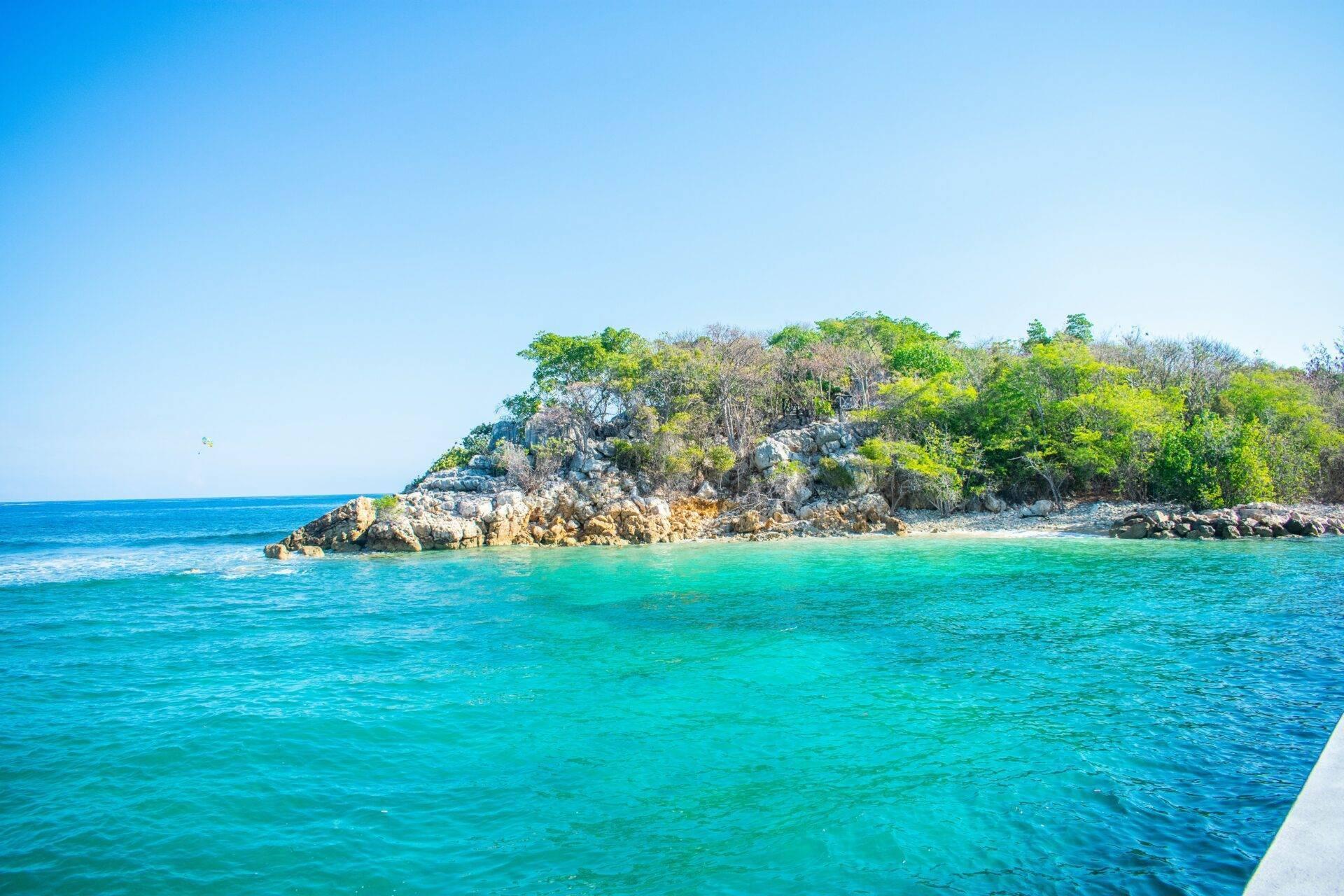 Une petite île rocheuse couverte de végétation tropicale émerge des eaux turquoise cristallines sous un ciel dégagé