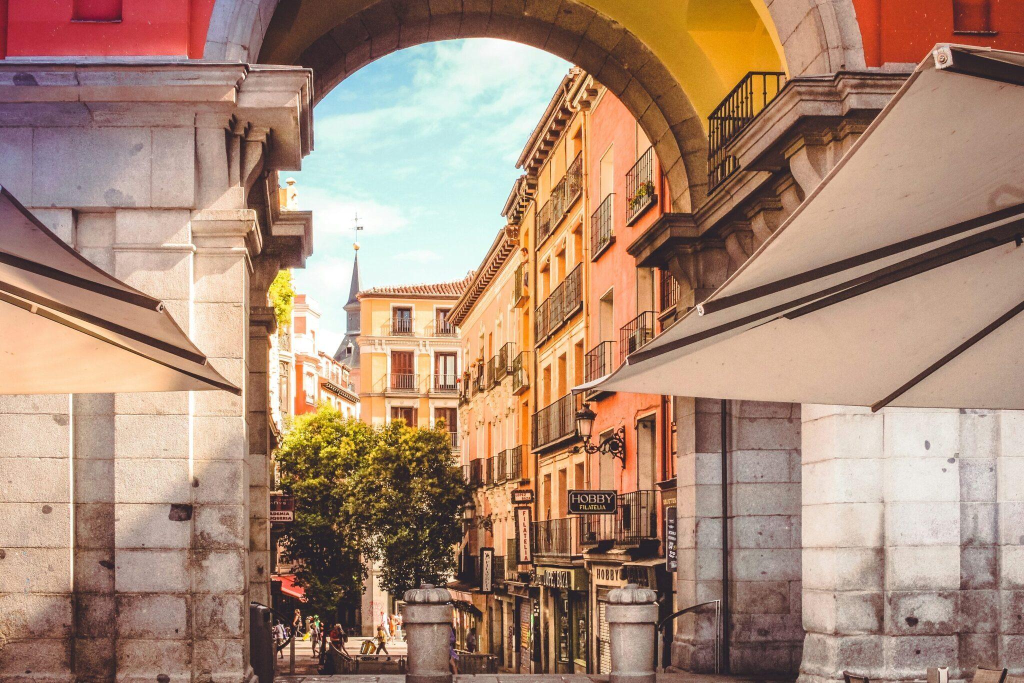 Una via storica di Madrid incorniciata da edifici arancioni e ocra con portici, insegne commerciali e cielo terso