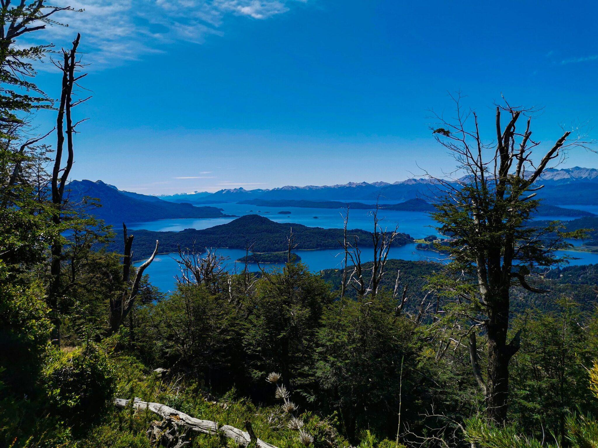 Panoramablick auf einen türkisfarbenen See, umgeben von dichten Wäldern und schneebedeckten Bergen unter klarem Himmel