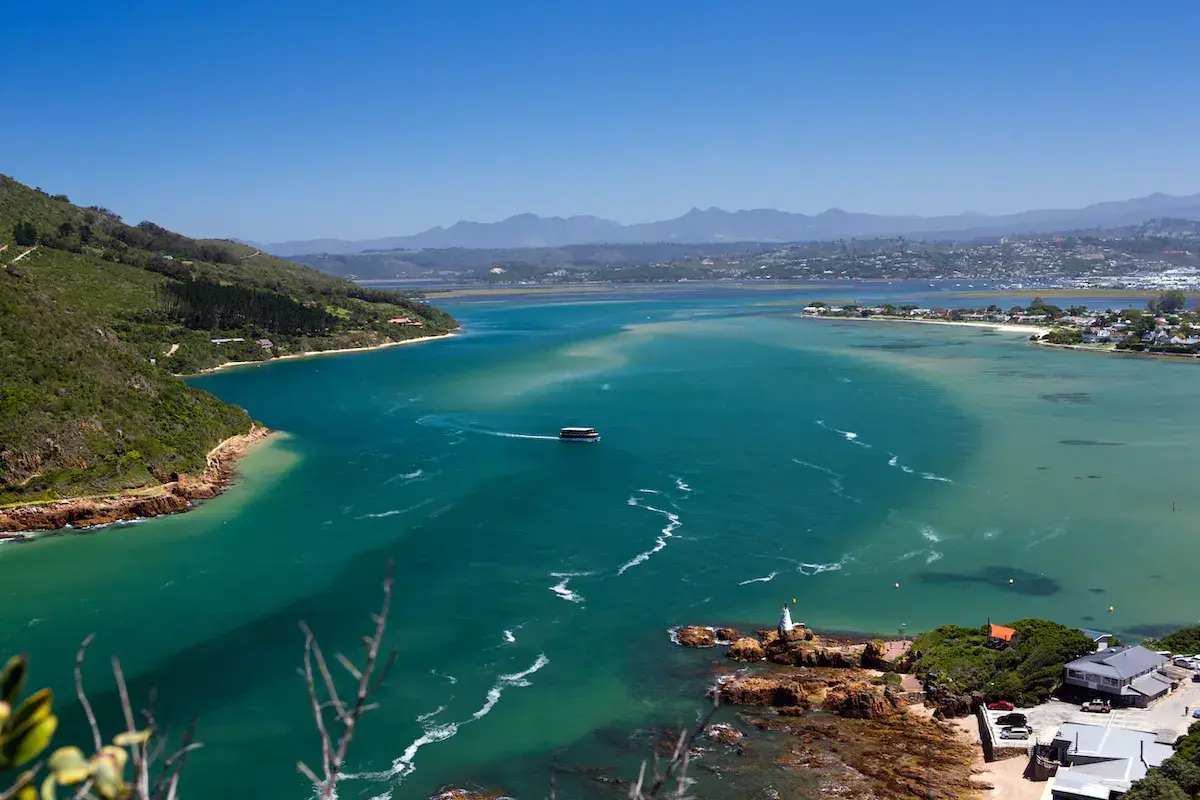 Vista aérea de una bahía costera con aguas turquesas, un pequeño faro, montañas al fondo y casas dispersas