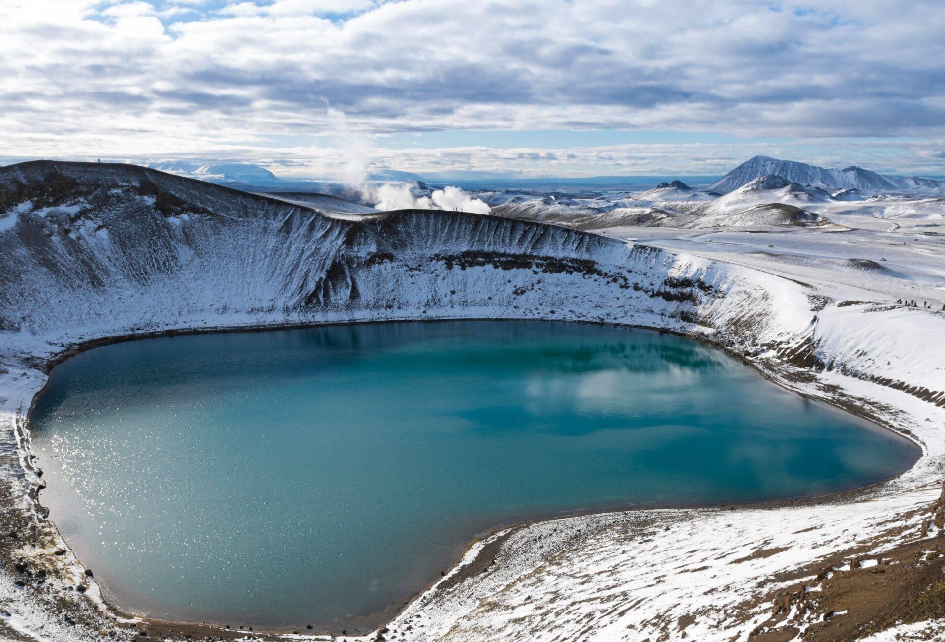 Un cráter volcánico nevado con un lago turquesa en su interior, rodeado de montañas nevadas bajo un cielo nublado
