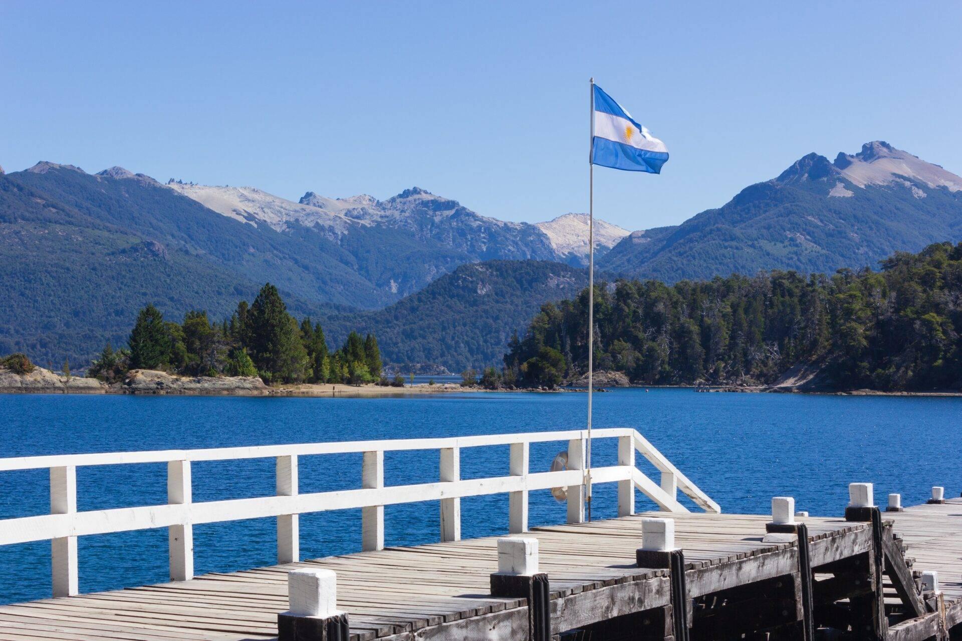 Un pontile in legno con bandiera argentina di fronte a un lago turchese circondato da montagne boscose