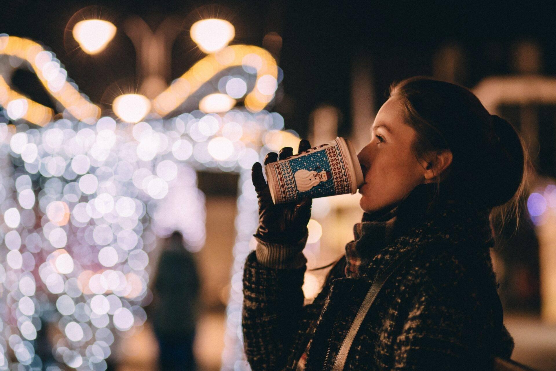 Una giovane donna beve una bevanda calda davanti alle luminarie di Natale scintillanti in città di notte