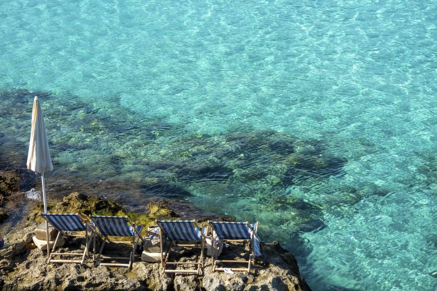 Gestreifte Liegestühle und weißer Sonnenschirm auf Felsen vor einem kristallklaren türkisfarbenen Mittelmeer