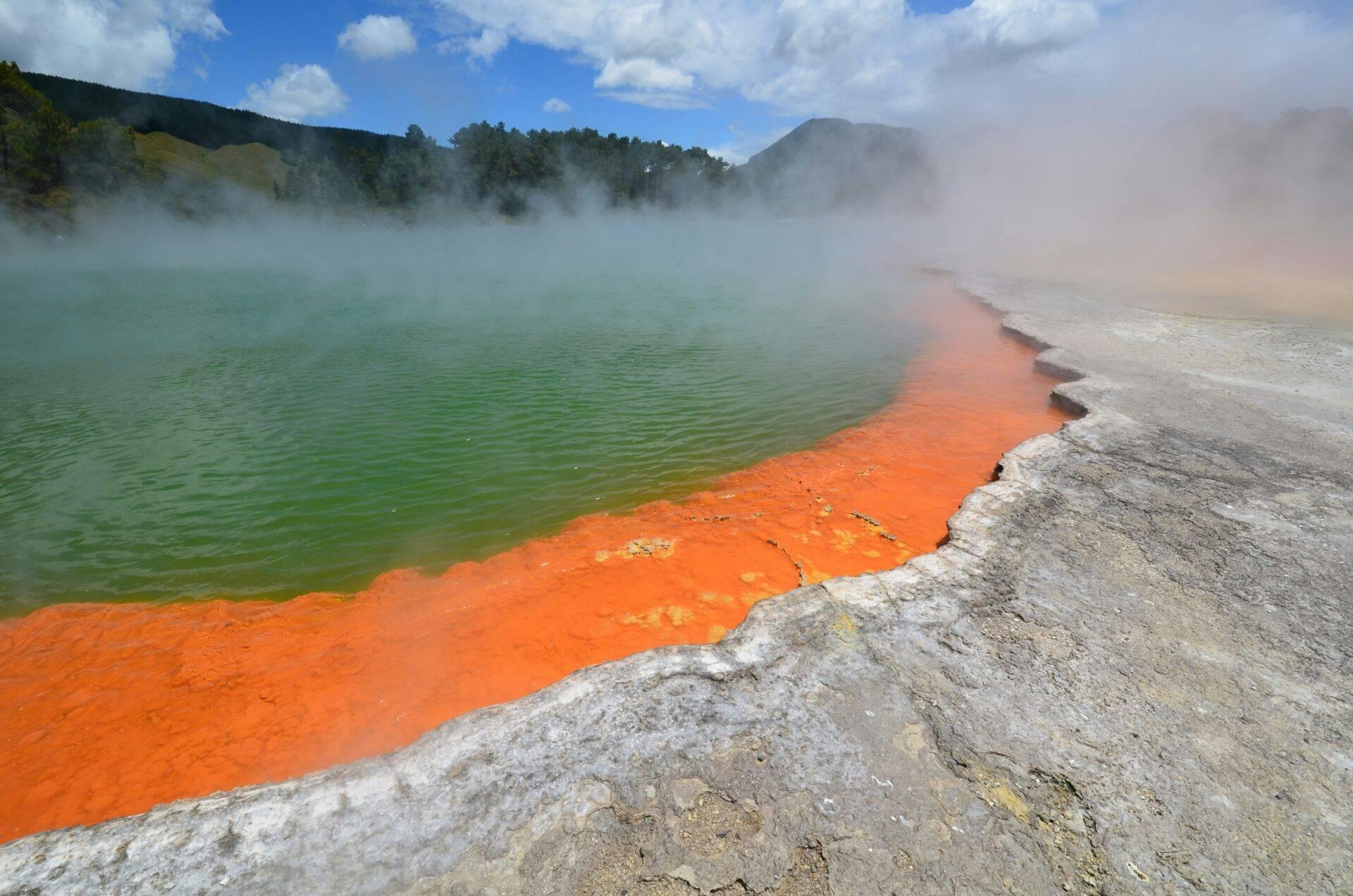 Un bacino geotermico con acque colorate verde smeraldo e arancione acceso, circondato da vapore e formazioni minerali bianche