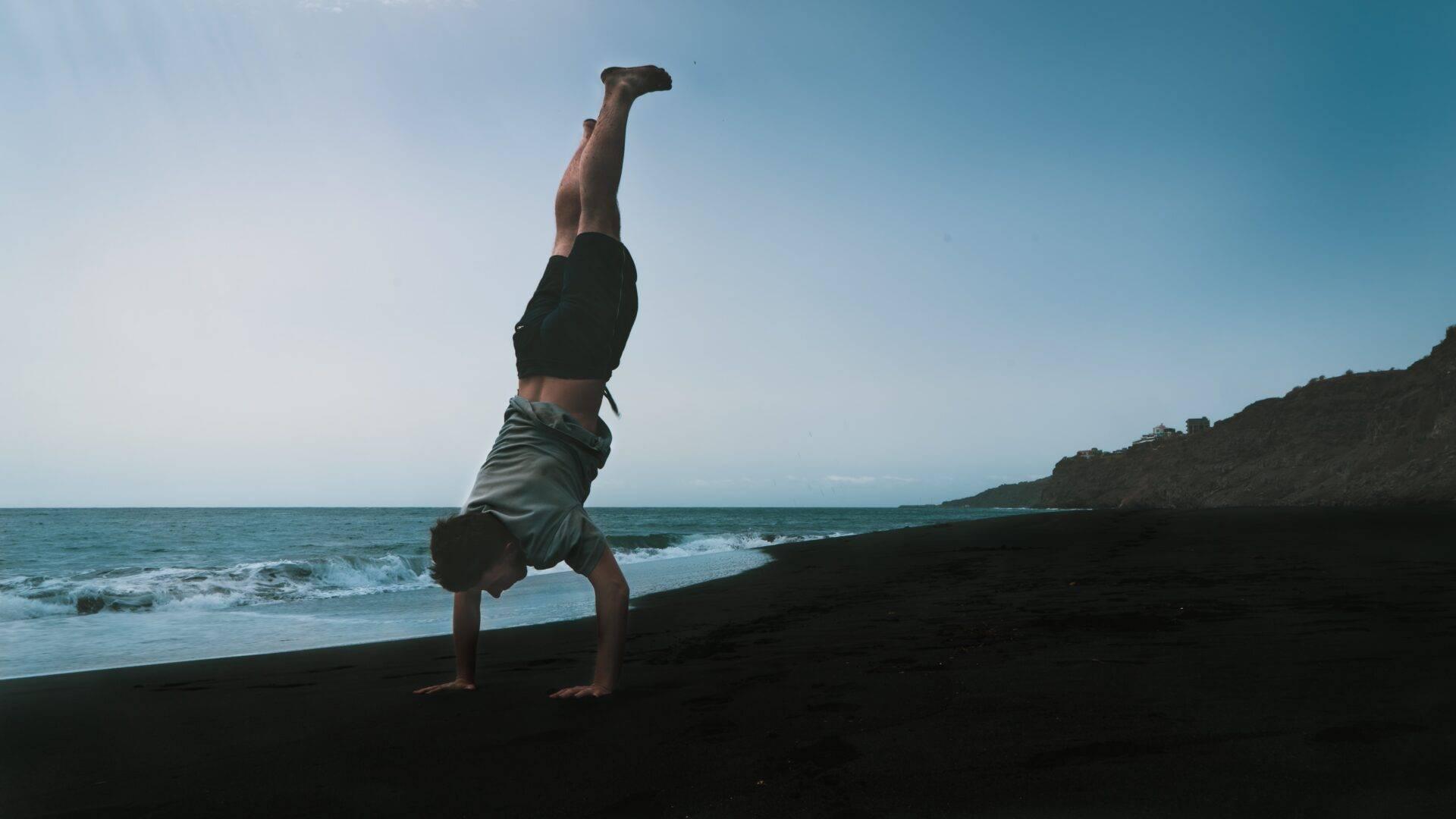 A man balancing on his hands on a black sand beach facing a turquoise ocean and cliffs